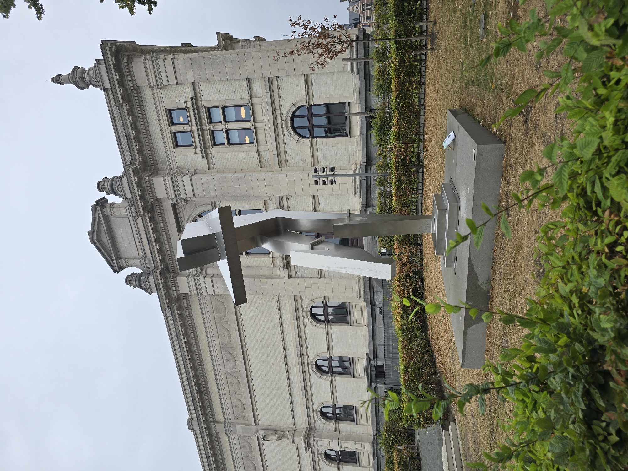 The image depicts a modern metal sculpture situated in front of a classical stone building with arched windows and ornate architectural details, indicative of a museum or public institution. The sculpture, with its abstract, angular form, is the focal point of the foreground, standing on a concrete base. Behind the sculpture, a neatly trimmed hedge forms a boundary along a dry, grassy area. The building itself is light-colored stone, with several stories and a grand facade featuring decorative carvings and pediments. The sky is overcast, suggesting a cloudy day. The overall scene is outdoors, likely in a public space or park adjacent to the building. Based on the architectural style of the building and the presence of notable public art, this location is likely the Royal Museum of Fine Arts Antwerp (KMSKA) in Antwerp, Belgium, a prominent cultural landmark.