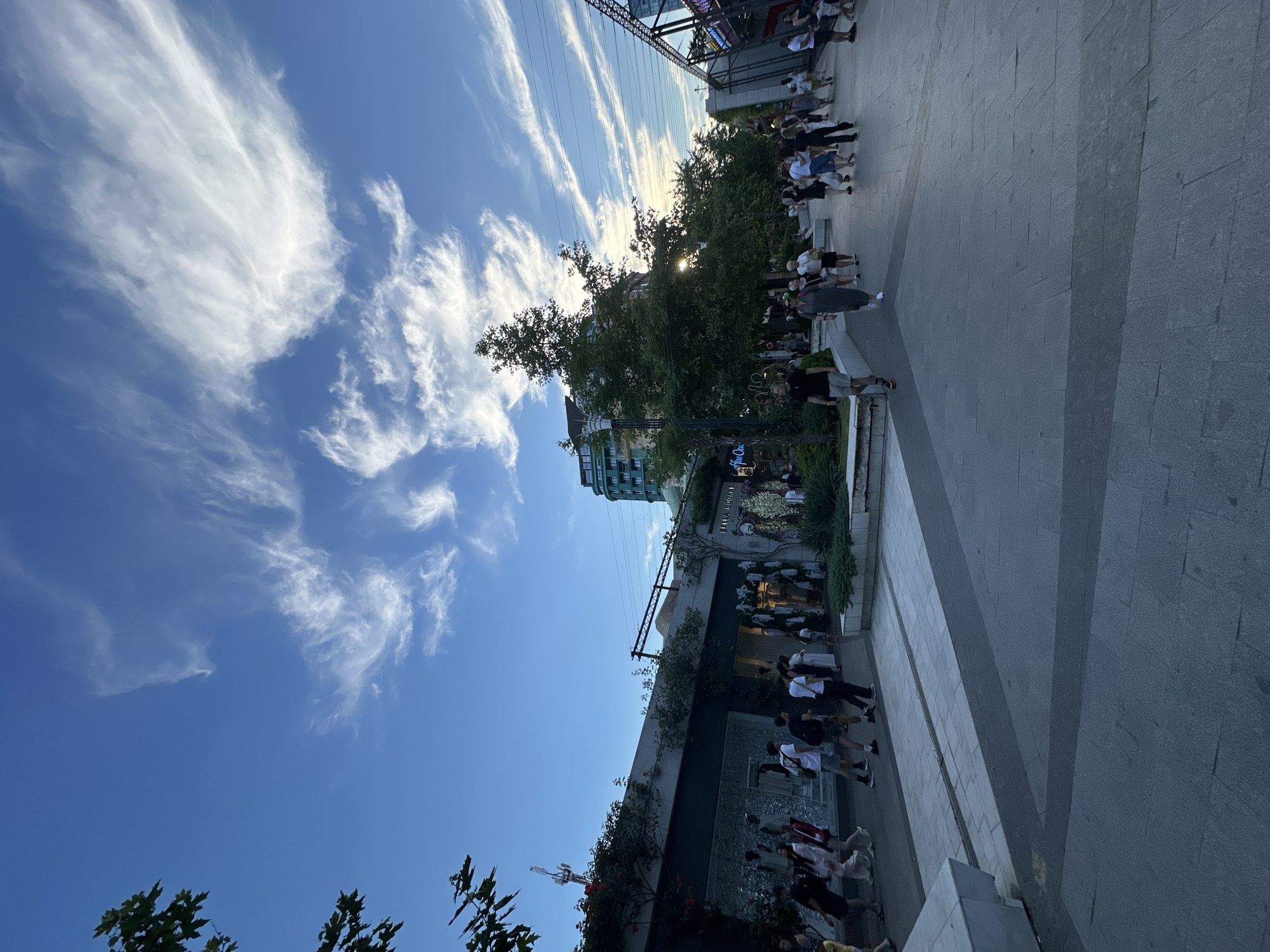 The image captures a lively outdoor scene with a clear blue sky streaked with wispy white clouds. Sunlight filters through the leaves of a large tree, casting dappled shadows on the ground. A paved walkway runs through the scene, lined with people in casual attire. Some individuals are walking, while others appear to be gathered, possibly interacting or observing something.
To the left of the walkway, there's a multi-story building with a modern architectural style. Lush greenery and plants are visible along the lower levels, suggesting a well-maintained urban or commercial area. The overall atmosphere seems to be one of a pleasant daytime gathering, perhaps a weekend or a leisurely afternoon. The sky's appearance suggests it's likely daytime, possibly late afternoon given the angle of the sun.
The arrangement of people and the presence of what looks like storefronts or cafes in the background hint at a public space, such as a pedestrian mall, a plaza, or a shopping district. The scene is devoid of any overt expressions of strong emotion, presenting a generally positive and active ambiance. No discernible text is clearly legible in the image.