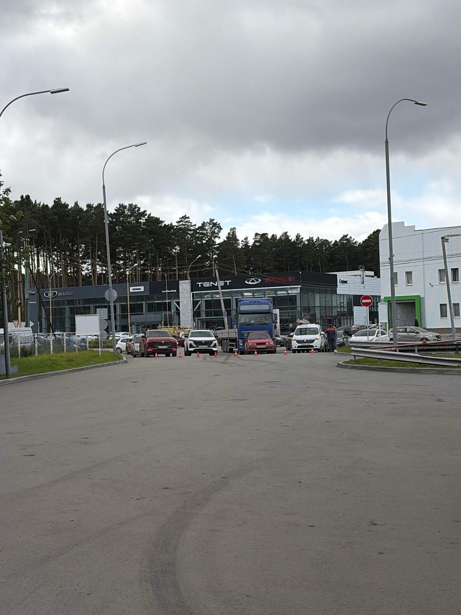The image shows an exterior view of a car dealership in Beryozovsky, Russia. The dealership has a modern facade with large windows and signs displaying brand names like "TENET" and "CHERY". In the foreground, there is an asphalt road with tire marks. Several cars are parked and driving in the scene, including a red SUV, a white van, and a blue truck. Traffic cones are placed on the road, possibly indicating a temporary obstruction or traffic control. A line of trees forms a backdrop to the dealership. The sky is cloudy, suggesting overcast weather.