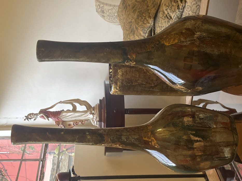 The scene is an interior residential space. In the foreground, two tall, reflective vases with slender necks and bulbous bases, dark in color with gold leaf accents, rest on a reflective tabletop. A third, shorter, cylindrical vase with corresponding decoration is partially visible between and behind them. A dark metal candlestick holding a red candle is partially visible to the left. A dark wooden console table with a single drawer in the midground supports a standing female figurine. The figurine, styled in an Art Deco manner, features a woman in a white gown with a red drape and an ornate headpiece. Behind the table, a window with reddish frames shows exterior greenery and a building structure. To the right, a segment of a sofa or armchair with patterned beige and gold upholstery and cushions is visible. No individuals are present. The specific location within Mexico City, Mexico, is indeterminable from the provided visual information.