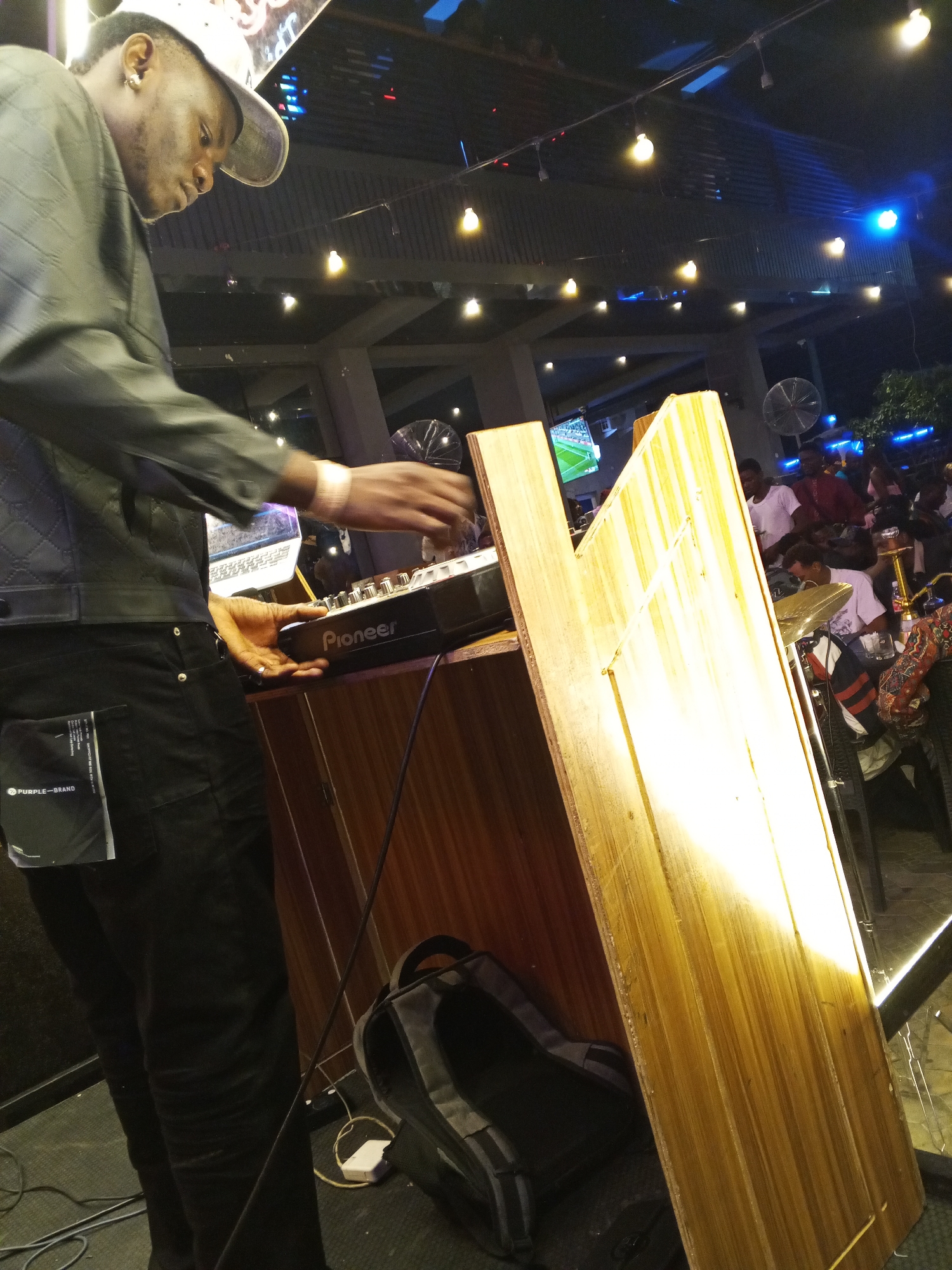 A person, likely a DJ, stands in a dimly lit indoor setting, possibly a bar or nightclub, in Lokoja, Nigeria. The DJ is operating a Pioneer mixing console mounted on a wooden DJ booth. The individual wears a backwards baseball cap, a dark jacket with quilted patterns, and black jeans with a label reading "PURPLE-BRAND" visible on the back pocket. A laptop is nearby. The background features a crowd of people, a television screen, and string lights.