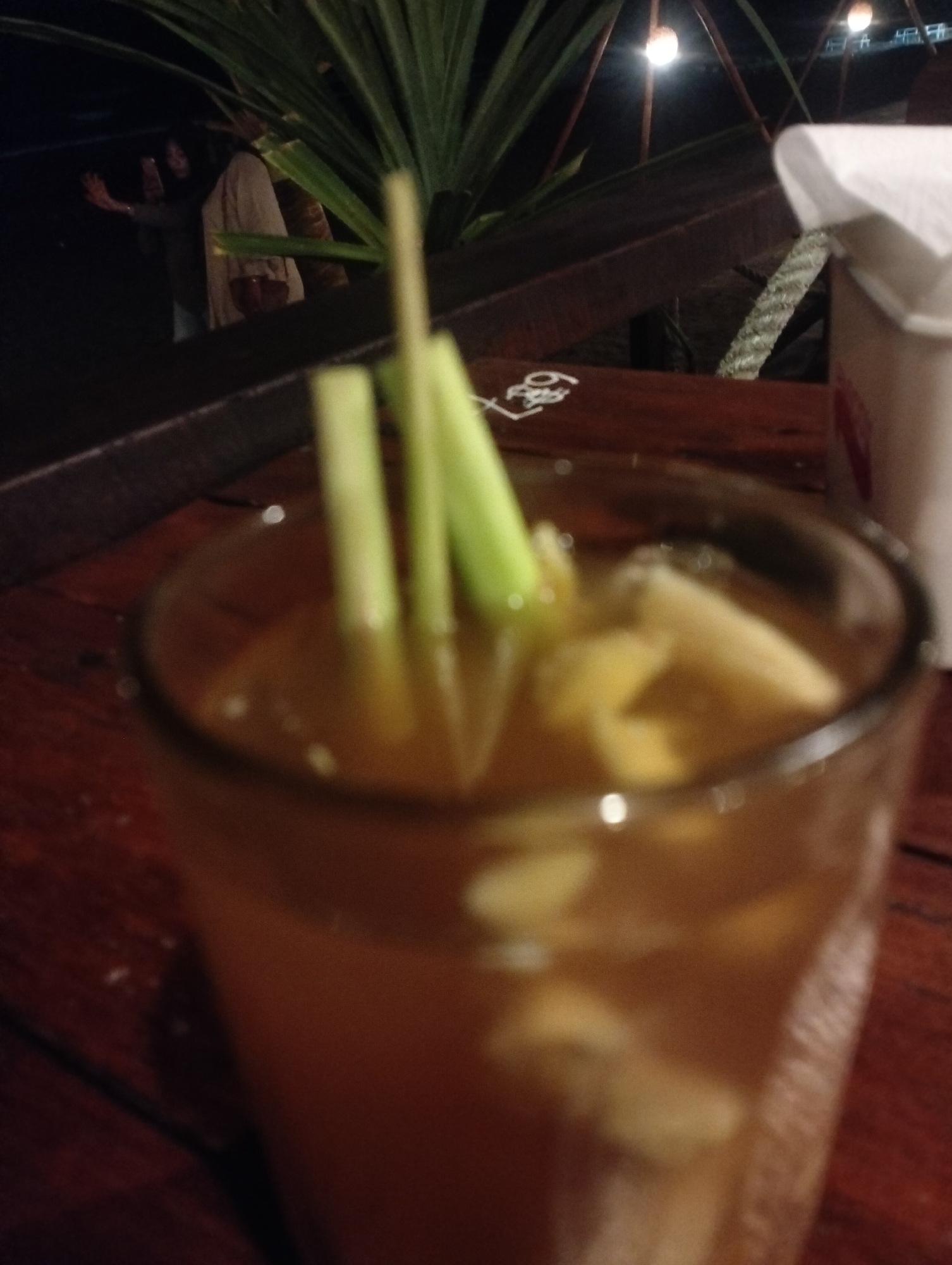 The image displays a close-up, slightly out-of-focus shot of a glass containing a light brown beverage, possibly a tea or herbal drink, situated on a dark wooden table. Inside the glass, several light-colored, irregular pieces, resembling ginger or cut fruit, are visible, along with two slender green stalks, likely lemongrass, standing upright as a garnish or ingredient.
To the right of the glass, on the same wooden table, a white paper napkin dispenser or cup is partially visible. Further back on the table surface, some faint white markings, including the number "28" and other indistinct symbols, are visible in one of the detailed crops.
The background is predominantly dark and blurry, indicating a nighttime setting or a dimly lit indoor environment. A large, leafy green plant, possibly a palm or similar tropical foliage, occupies the upper left portion of the background. Beyond the plant, a person is faintly discernible in the distance, appearing to hold a device or gesture with an arm. Several small, glowing artificial lights, resembling bare bulbs or decorative lights, can be seen in the far background, suggesting an overhead structure or canopy. The scene evokes a casual evening atmosphere, likely at an outdoor or semi-outdoor cafe or restaurant in Keudekruenggeukueh, Indonesia. The primary focus is on the prepared drink.