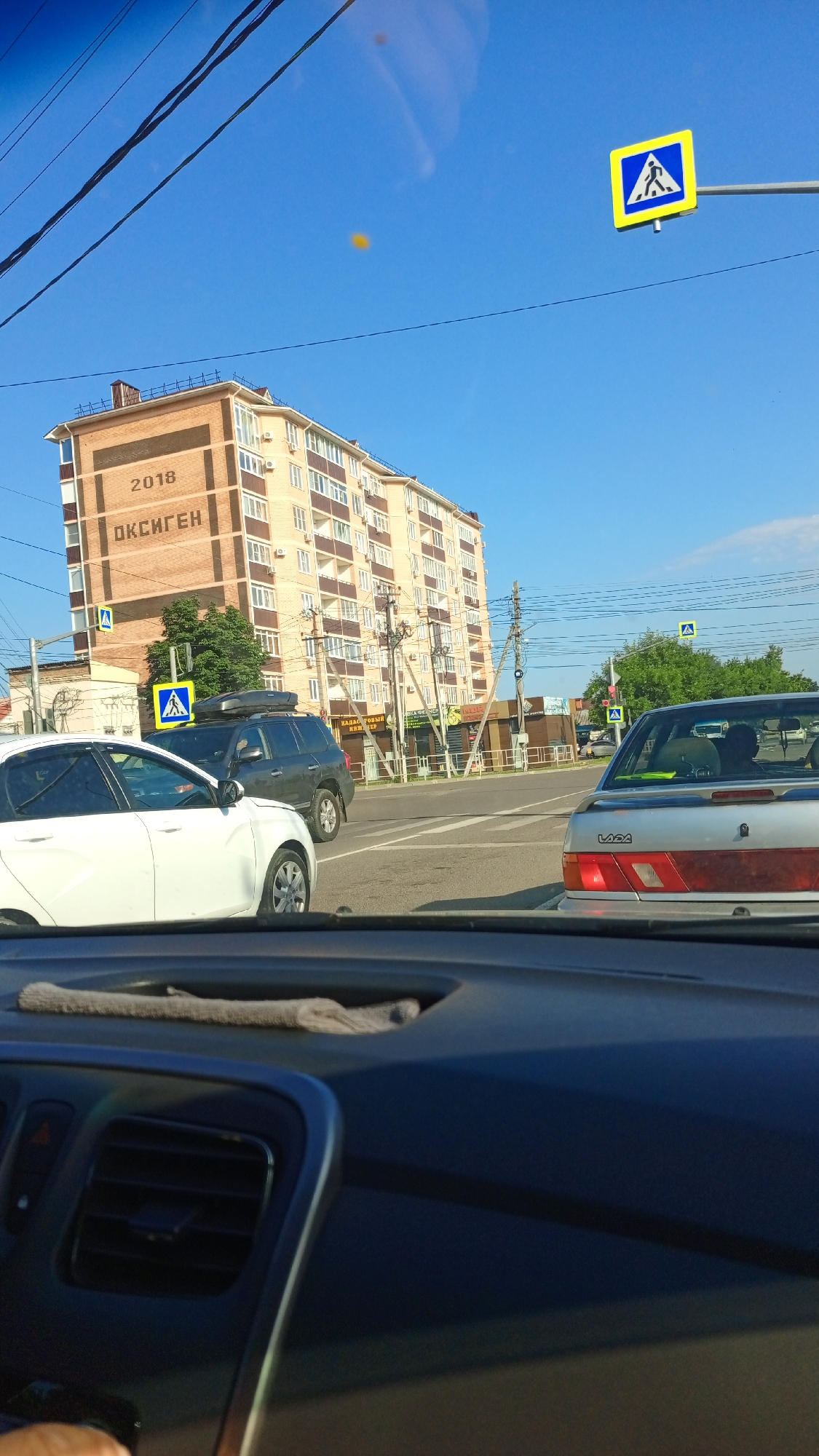 In Belorechensk, Russia, on a clear day, a view from inside a vehicle shows a multi-story building with the numbers "2018" and the word "ОКСИГЕН" (Oxygen) written on its facade. Traffic signs indicating a pedestrian crossing are visible, one on a pole to the right and another lower down on the left. Cars are stopped at an intersection, including a white car in the foreground, a dark SUV with a roof rack, and a silver Lada sedan. The urban environment includes power lines and trees, with storefronts visible in the background. The interior of the vehicle shows part of the dashboard and windshield, with a cloth draped over the dashboard.