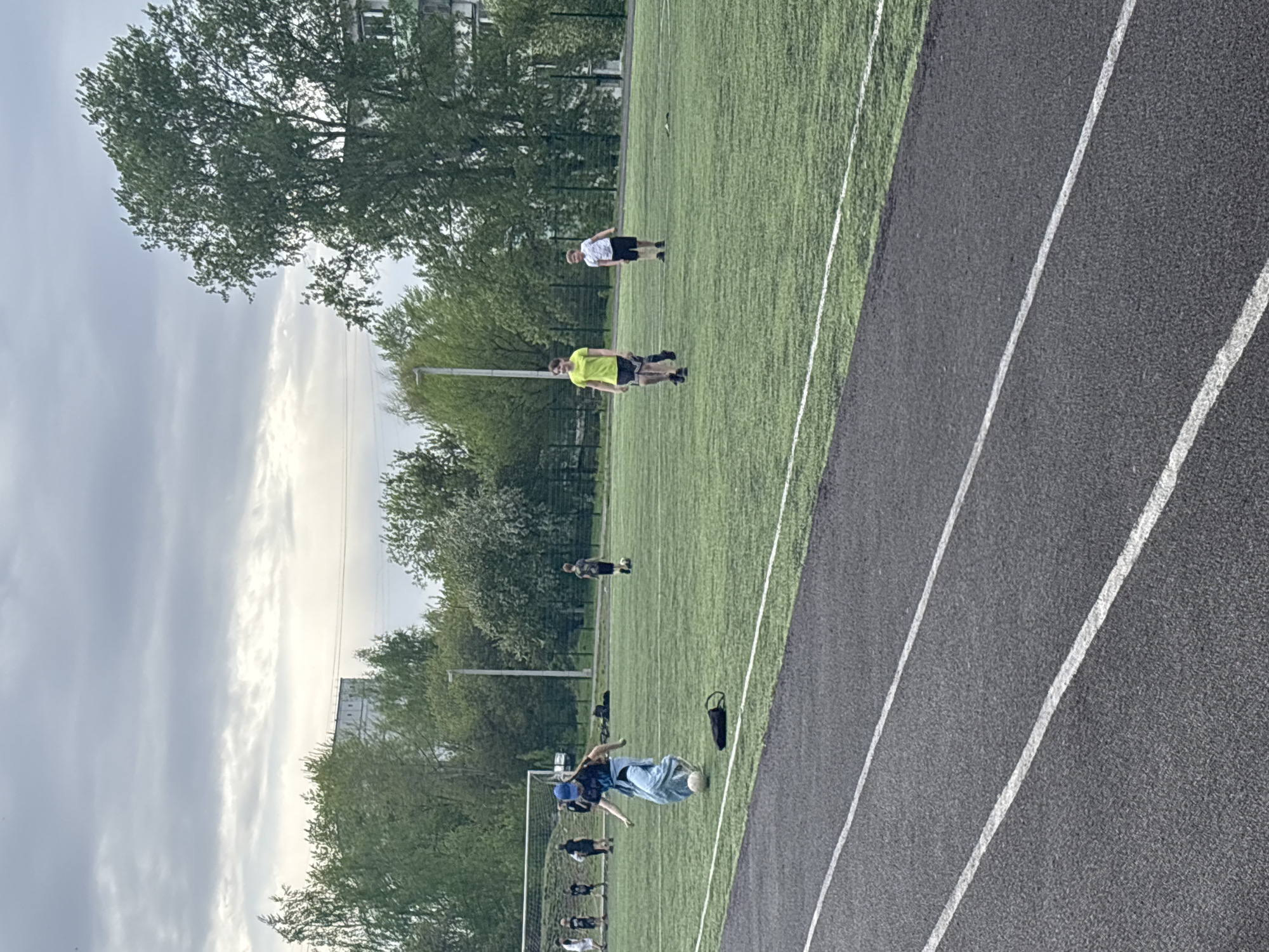The image shows an outdoor scene on what appears to be a sports field. There are people scattered around the field, some near a soccer goal. The field is green, possibly artificial turf, and is bordered by a running track with white lane markers. Trees surround the perimeter of the field and a building can be seen in the background on the left. Based on the prompt, this location is in the city of Chaikovsky, Russia. People are present on the field, possibly playing a sport or engaged in some training activity. There is an object, potentially a bag, placed near the track. One individual is performing a kicking motion. The sky appears overcast.