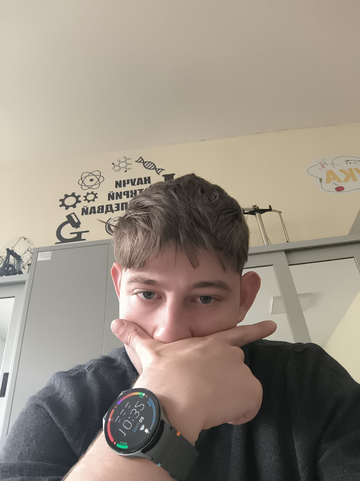 A young man is posing in a selfie, with a curious expression. He has short brown hair and blue eyes. He is wearing a black sweater and a smartwatch. The time on the watch reads 10:35. The background is a wall with scientific drawings of a microscope, atom model, molecule, DNA helix, gear. Some words are written in Bulgarian. The drawing of a happy face with a cherry nose, is also visible. Behind the man, there is a grey cabinet. Given the writing on the wall, it is likely that the photo was taken in a classroom. I cannot know the exact location of the photo within Sofia.