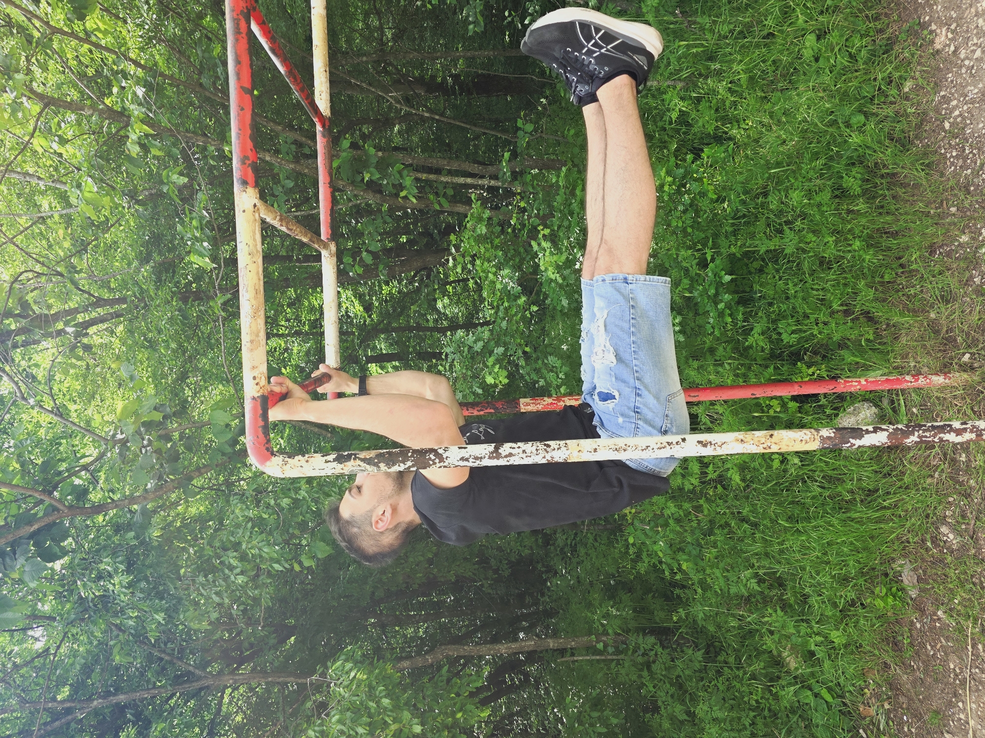 A man is hanging upside down from a red and white metal structure. He is wearing a black t-shirt, denim shorts, and black shoes. The structure is surrounded by green foliage, which appears to be trees and grass. The scene takes place in the city of Shumen, Bulgaria.