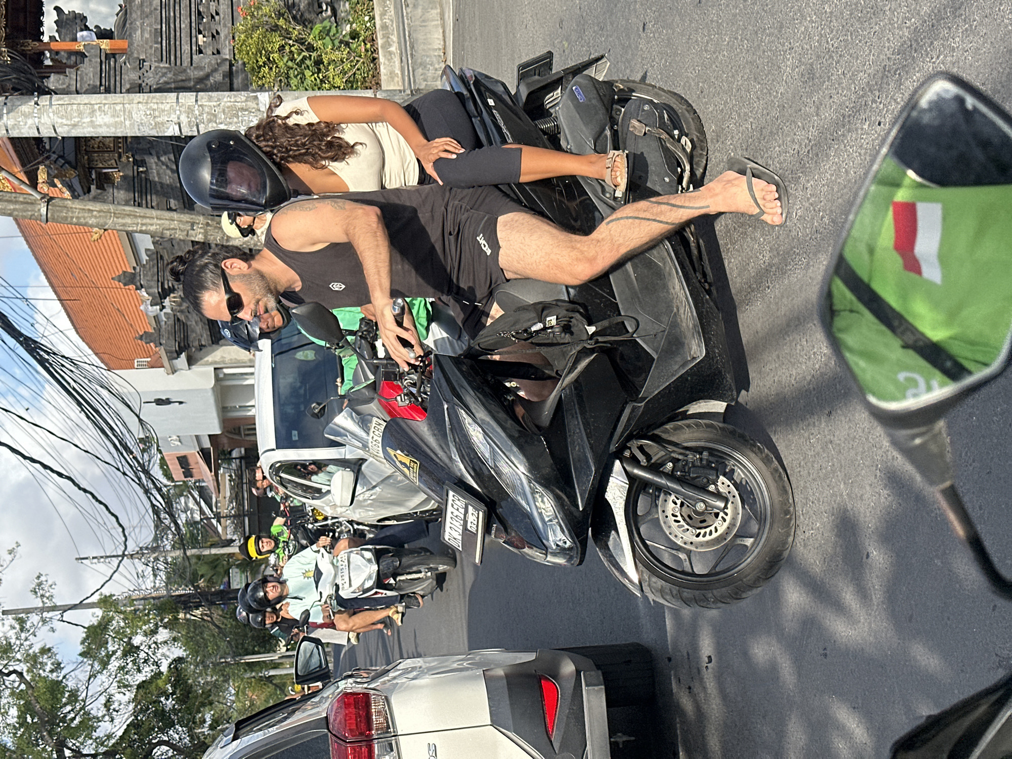 The image shows a busy street scene in North Kuta, Indonesia, likely during daylight hours. In the foreground, a black scooter is prominent, with a man and a woman riding on it. The man is driving, wearing a black tank top, black shorts, sunglasses, and flip-flops. He has a bun in his hair and a distinctive tattoo on his leg. The woman is the passenger, wearing a light-colored top and dark leggings, with long curly hair and a black helmet. She is wearing sandals.
Behind them, other motorcycles and a white car are visible, indicating traffic congestion. The architecture in the background suggests a tropical urban environment, with buildings displaying traditional Indonesian motifs, such as ornate stonework and tiled roofs. Overhead, a dense network of electrical wires crisscrosses the sky, a common sight in many Indonesian cities. The road surface is asphalt, and shadows of objects and trees are cast on it, suggesting a sunny day. The overall impression is one of movement and daily life in a bustling Indonesian city.