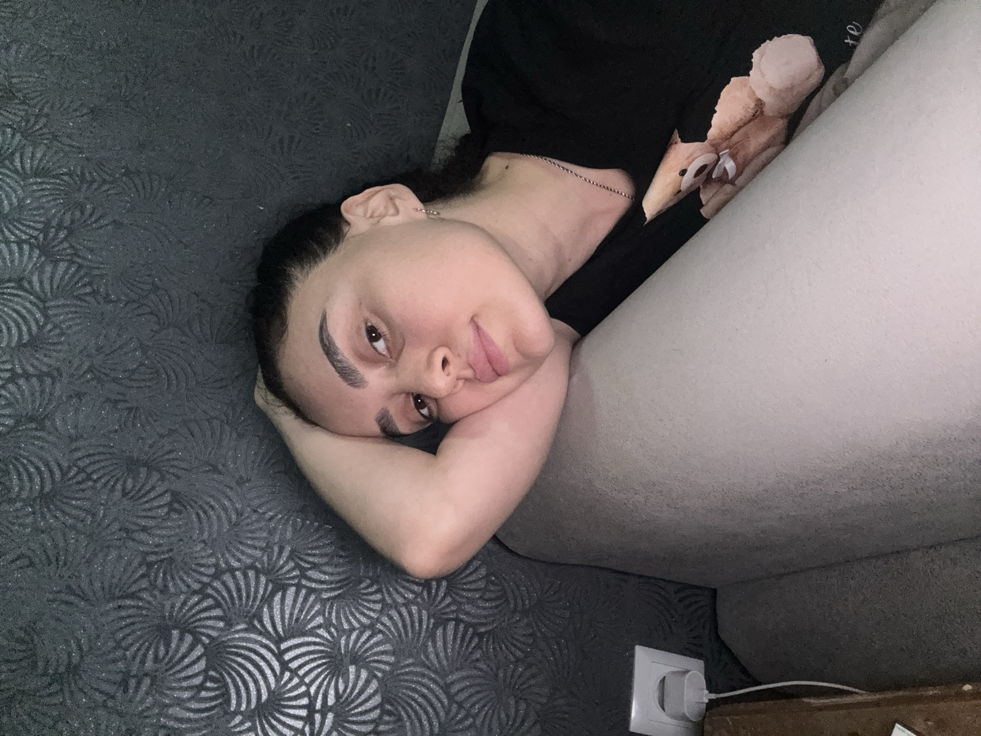 A woman is lying on a patterned gray surface, resting her head on her arm. She is partially reclined against a white upholstered object, possibly a sofa or cushion. She is wearing a black top with a graphic on the chest, and a thin chain necklace is visible. Her hair is dark and pulled back. In the lower right corner of the image is a white electrical outlet with a charger plugged in. The scene appears to be indoors, possibly in a residential setting, but without more context, it is impossible to determine if this photo was taken in Tbilisi, Georgia.