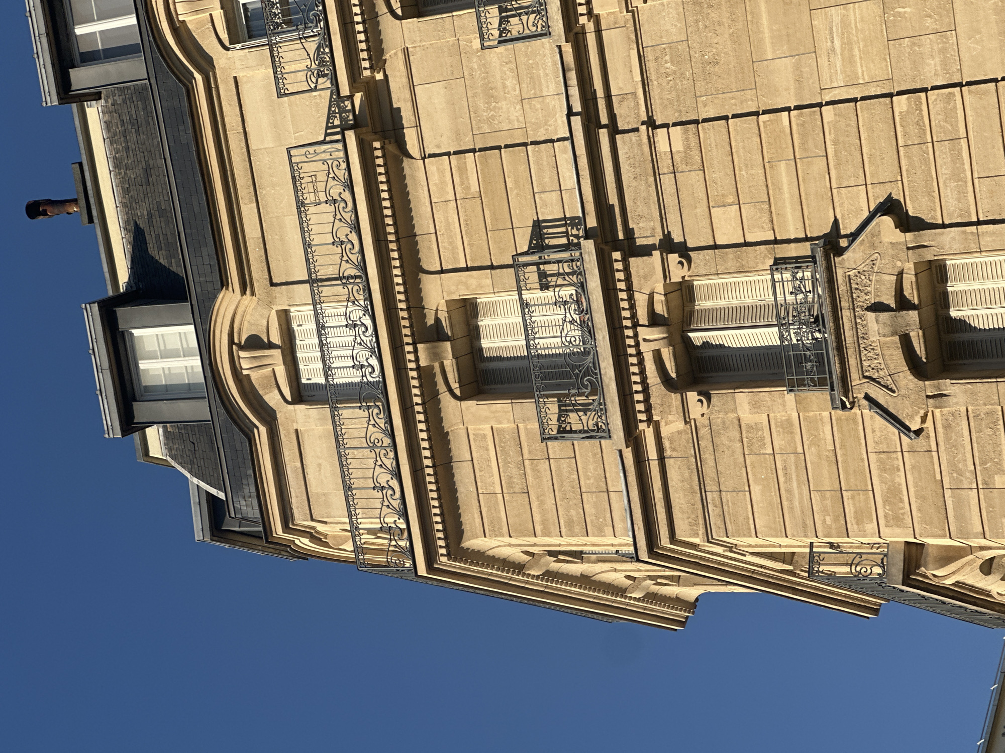 The image shows a close-up, eye-level view of the upper floors of a Parisian Haussmann-style building, bathed in warm, late afternoon sunlight. The building's facade is constructed of light-colored stone blocks, intricately detailed with pilasters, cornices, and decorative moldings around the windows. Several balconies with ornate wrought-iron railings punctuate the facade, each adorned with intricate scrollwork. The windows are closed, fitted with white louvered shutters, and set within carved stone surrounds. Above the main facade, a dark slate mansard roof is visible, punctuated by dormer windows with similar white shutters. A single, weathered, reddish-brown chimney pipe extends from the roofline. The clear, deep blue sky occupies the left portion of the frame, contrasting with the warm tones of the building. The scene captures the architectural elegance typical of the Haussmannian style found throughout Paris.