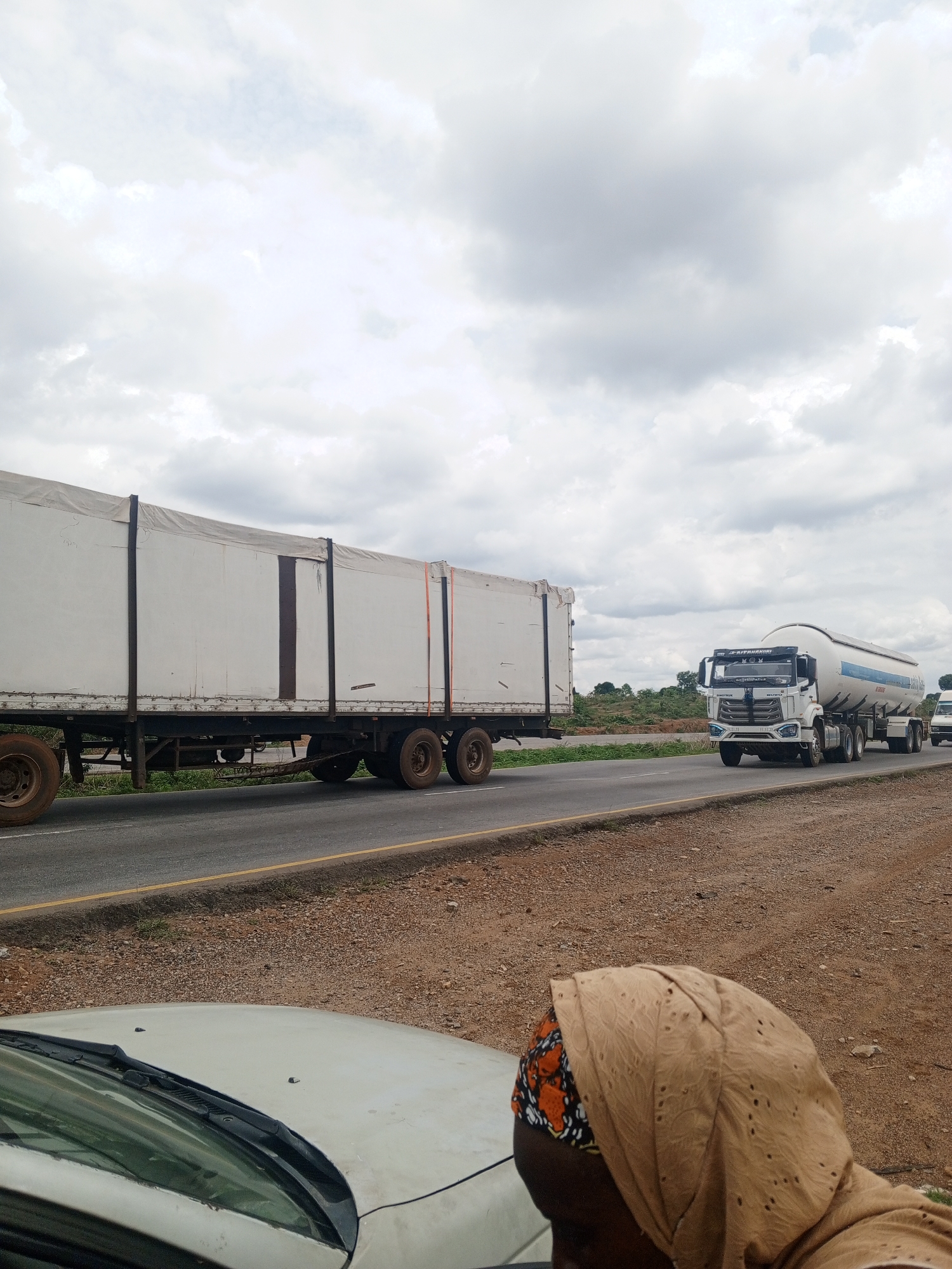 The image shows a scene on a highway in an unidentified location. A large white trailer truck is traveling on the road, followed by a white tanker truck with a rounded tank. The sky above is overcast with gray clouds. In the foreground, the hood of a car is visible, along with a person wearing a brown head covering with an orange and black patterned edge. The area beside the road is brown and appears to be dirt or gravel.