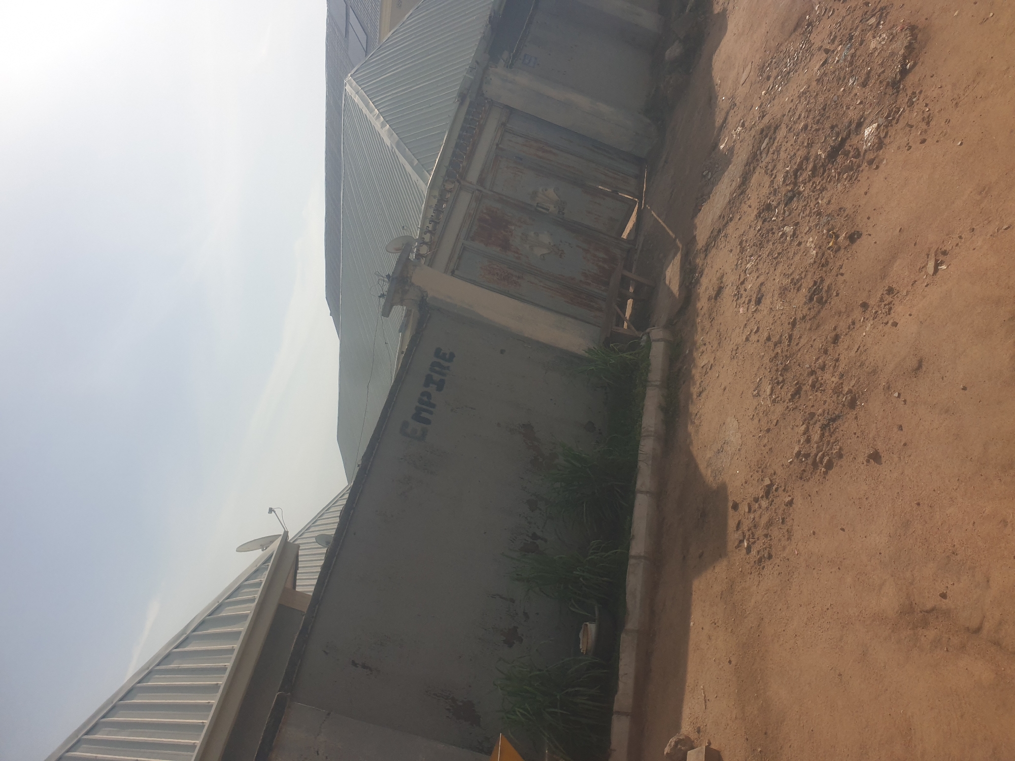 In Jauro Yinu, Nigeria, a weathered concrete wall, painted grey, stands prominently in the foreground, displaying signs of age and decay with patches of darker discoloration. The word "EMPIRE" is spray-painted in dark blue lettering across its surface. To the right of the wall is a rusty, double-doored metal gate, flanked by concrete pillars and a short section of barbed wire fence atop the wall. Green grass grows along the base of the wall in front of a concrete kerb. The ground in front of the wall and gate is a mixture of reddish-brown earth, scattered small stones, and debris. In the background, a building with a corrugated metal roof is visible, along with portions of another structure. The sky is bright and hazy, suggesting daylight.