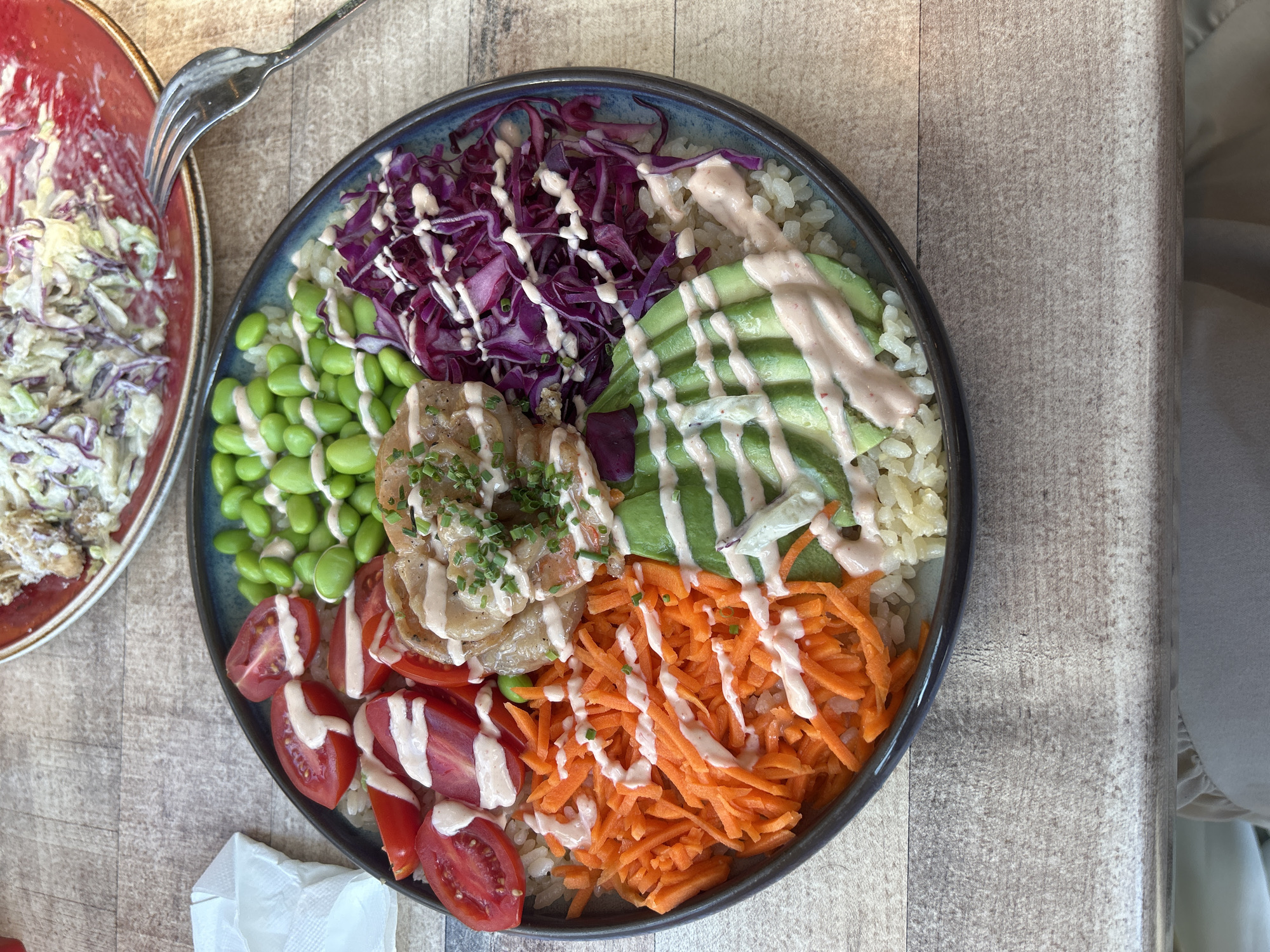 A large, round, dark-rimmed bowl containing a vibrant, segmented meal is centrally positioned on a light-colored wooden table, allegedly located in Sofia, Bulgaria. The meal consists of a base of light-colored grains, topped with distinct sections arranged radially: sliced red tomatoes, shelled green edamame, cooked protein (likely shrimp or chicken) garnished with chopped green chives, shredded purple cabbage, fanned slices of avocado, and shredded orange carrots. A light-colored, creamy sauce is drizzled over the tomatoes, edamame, protein, cabbage, and carrots. A reddish-pink sauce is drizzled over the avocado. In the upper right background, a partial view of a red ceramic plate is visible, holding a salad or coleslaw mix with green and purple cabbage and a creamy dressing. A silver fork, displaying residue from the salad, rests on the red plate and the wooden table. A crumpled white paper napkin is situated on the table in the upper left. A portion of light-colored fabric is visible beneath the table in the lower background, indicating the presence of a seated individual.