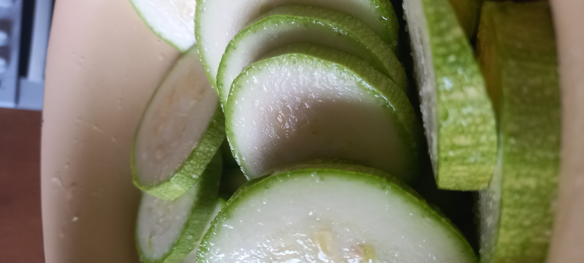 The image presents a close-up, vertical shot of numerous round slices of a green-skinned vegetable, likely zucchini or squash, arranged within a light beige or cream-colored container. Each slice, approximately 0.5 to 1 cm thick, features a pale green rind and a lighter, off-white to yellowish-white fleshy interior, where small, pale seeds or remnants are faintly visible. The surfaces of the vegetable slices are glistening with moisture, suggesting they are wet, possibly from washing or light salting, as small white particulate matter, consistent with salt crystals, adheres to the rinds and cut surfaces of several slices.
The slices are stacked and overlapped, creating depth, with some lying flat in the foreground and others standing partially upright in the mid-ground. The container, made of a smooth, opaque material, occupies the lower portion and edges of the frame. The background is out of focus, showing a blurred reddish-brown surface on the upper left and a blurred light grey or white object, possibly a kitchen appliance or fixture, on the upper right. No individuals, specific actions, or environmental details that would allow for geographical identification, such as the city of Kryvyi Rih, Ukraine, are present within the visual information provided.