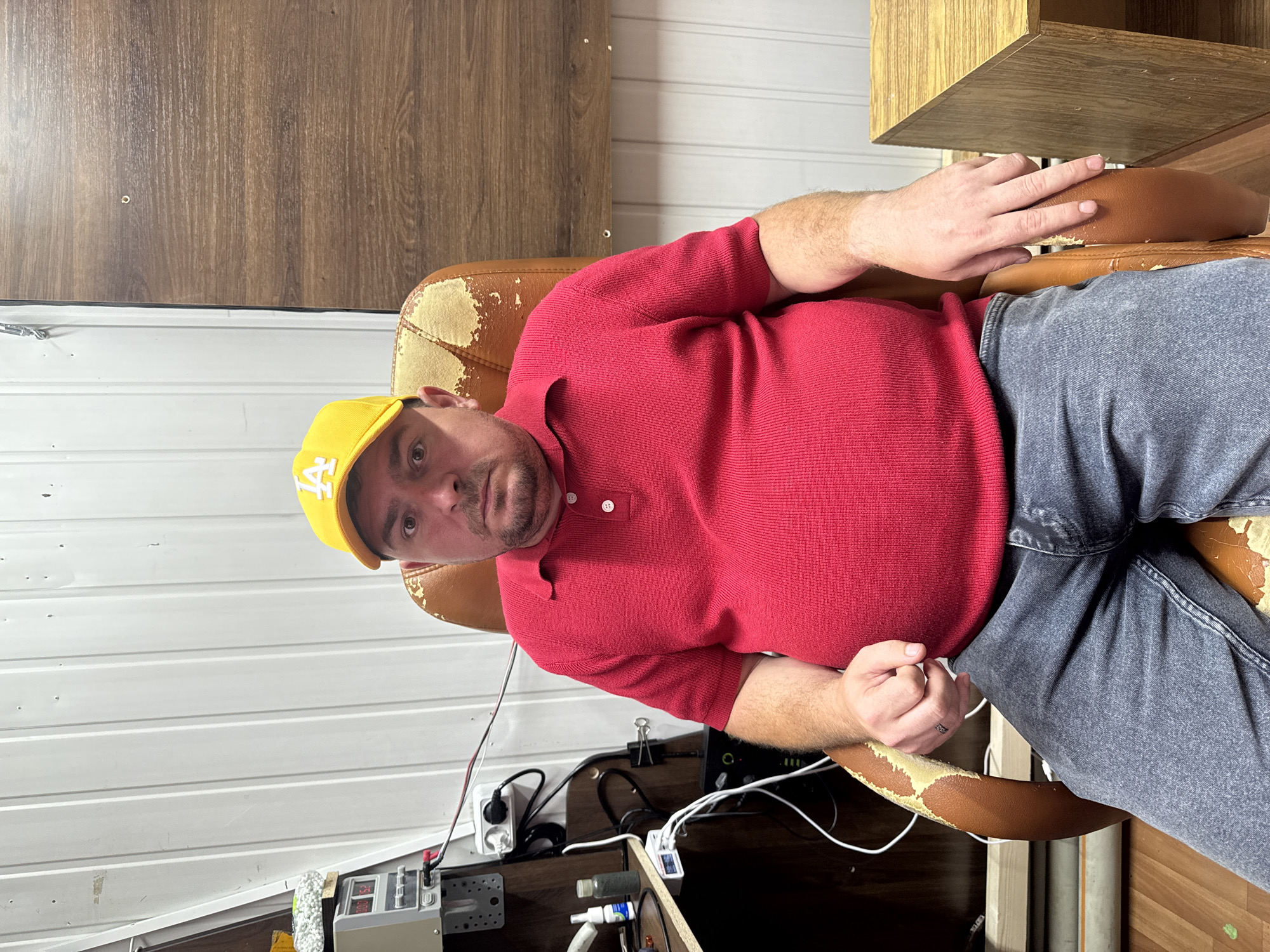 A man wearing a yellow cap and a red polo shirt is sitting in a worn leather office chair. He is looking directly at the camera with a neutral expression. The background consists of a white, corrugated metal wall and a wooden panel. To the left of the man, there is a desk with electronic equipment and cables. To the right, a portion of a wooden table is visible. The overall scene appears to be an indoor workspace or utility area.