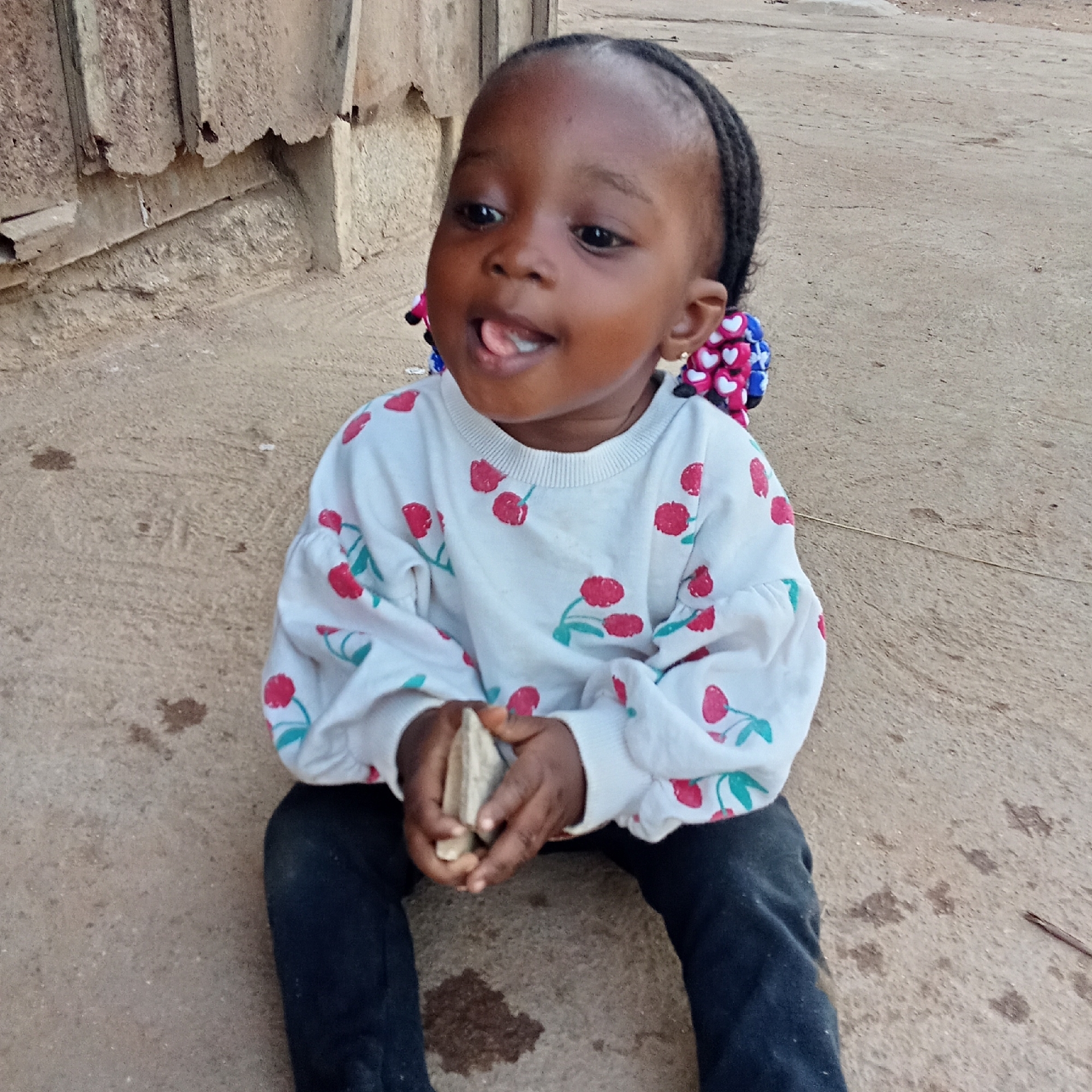 A young, dark-skinned child is seated on the ground outdoors, looking upwards and to the left with a playful expression, her tongue slightly out. She is wearing a white long-sleeved shirt with red cherry patterns and black pants. Her hair is braided and adorned with colorful accessories featuring blue, pink, and white heart shapes. In her hands, she holds two light-colored, natural objects, possibly small stones or pieces of wood.
The setting appears to be a casual outdoor area during daytime, with natural light. The ground is a mix of concrete and dirt, showing some marks and debris. Behind the child, there is a rustic, weathered wall or structure, possibly made of concrete or corrugated metal, exhibiting signs of age and wear. The scene suggests a candid moment of the child playing outdoors in a simple environment, aligning with the provided location context of Akwanga, Nigeria. There is no visible text in the image.