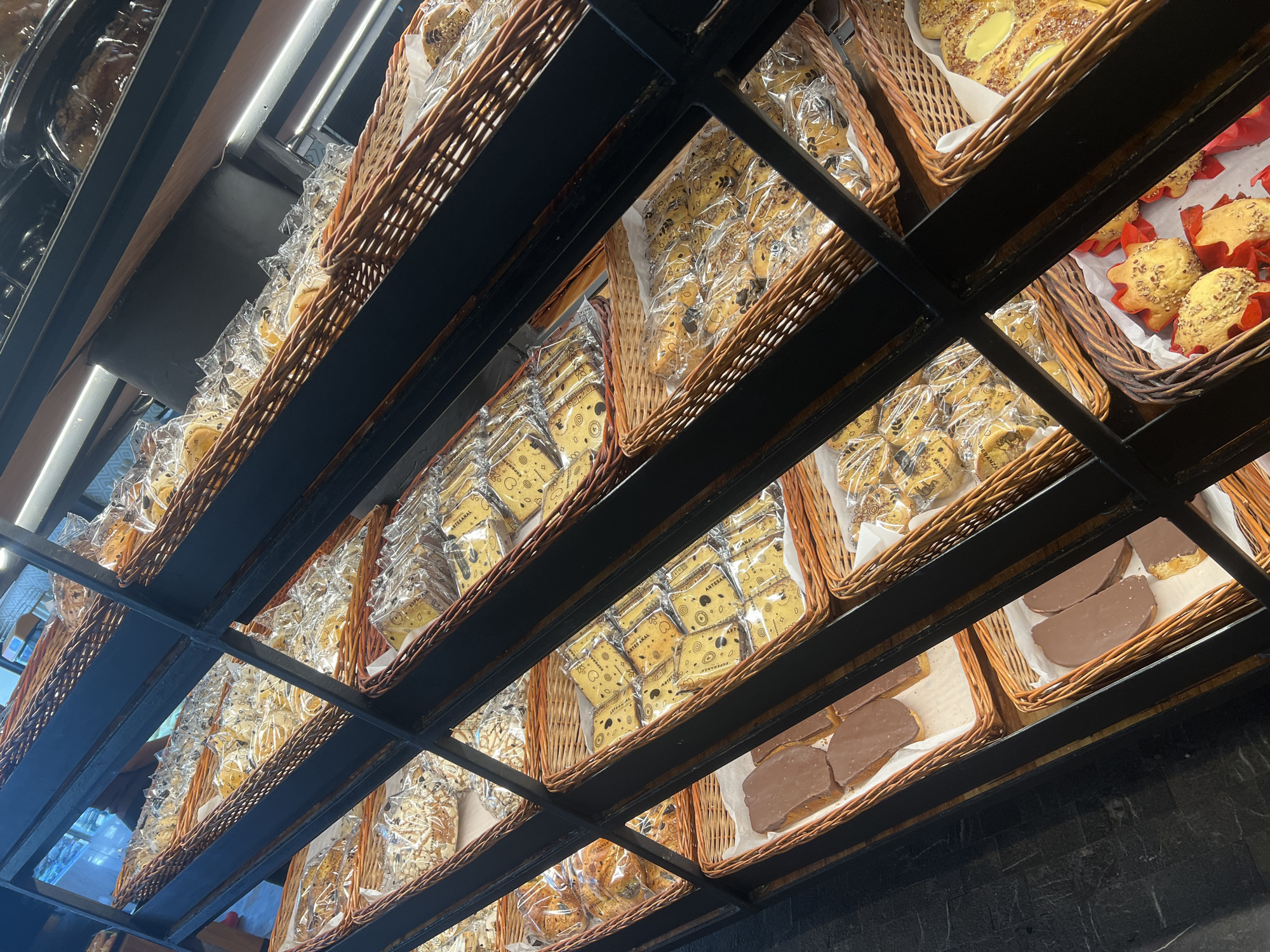 The image shows a low-angle shot of shelves filled with wicker baskets containing baked goods, possibly in Mexico City, Mexico. The shelves are organized into a black metal grid structure, with the baskets neatly arranged. The baked goods include pastries wrapped in clear plastic packaging, some labeled "Artesanal" and "Esperanza", along with other unpackaged items like chocolate-covered pastries and muffins in red paper cups. The lighting is bright, illuminating the products on display. The view is from below, looking upwards towards the shelves.