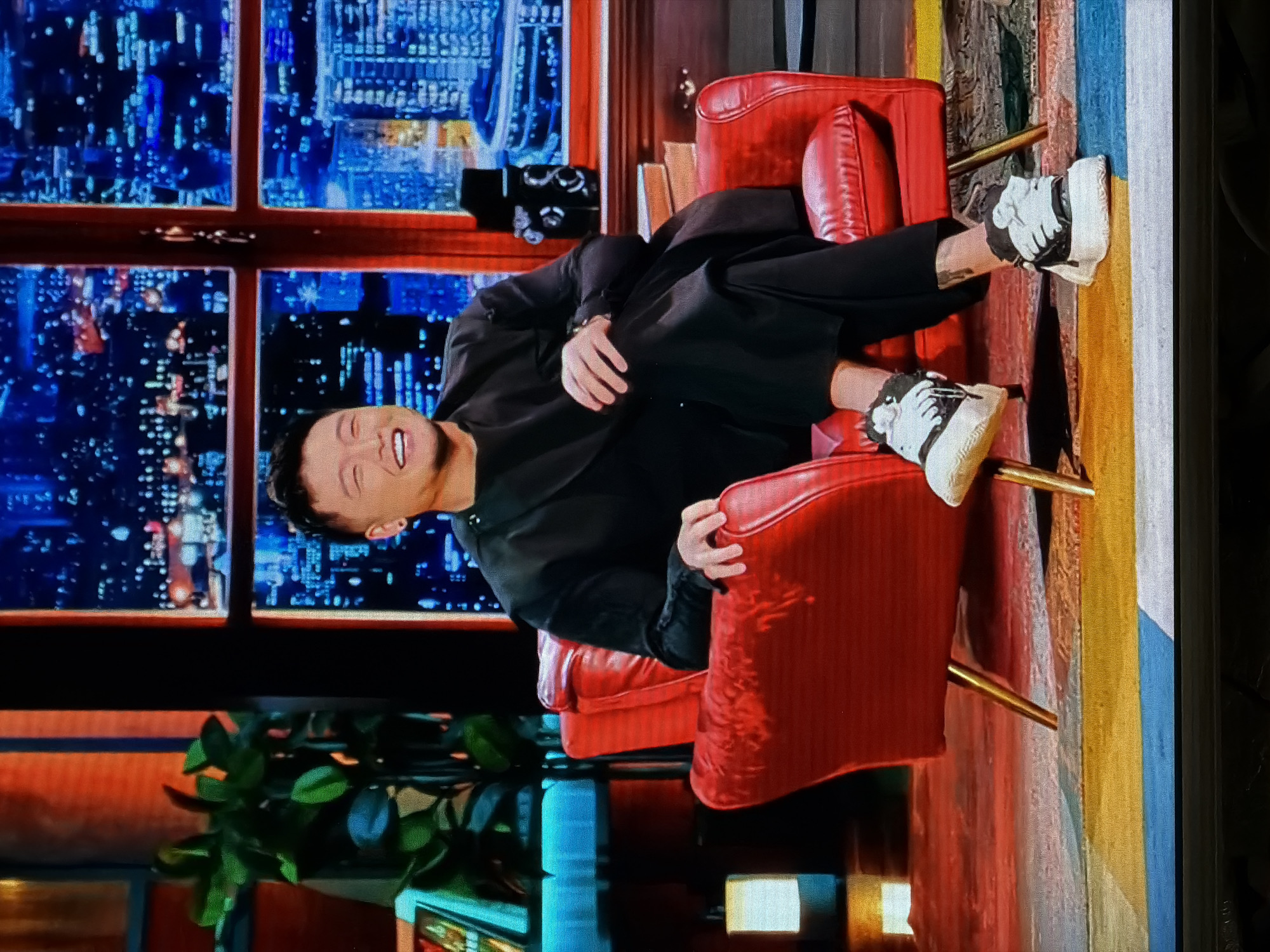 A man in a black suit and black pants sits in a red leather chair with gold legs, facing the viewer. He is smiling. He is wearing white sneakers with black accents. Behind him is a window showing a cityscape at night, likely depicting Moscow, Russia. The cityscape is dominated by high-rise buildings with many illuminated windows. A green plant is visible in the lower-left corner of the image. There are books and a decorative object on the windowsill behind the man. A tapestry with gold and blue detailing hangs beside the chair. There is a light reflecting from the bottom. The image has been rotated.