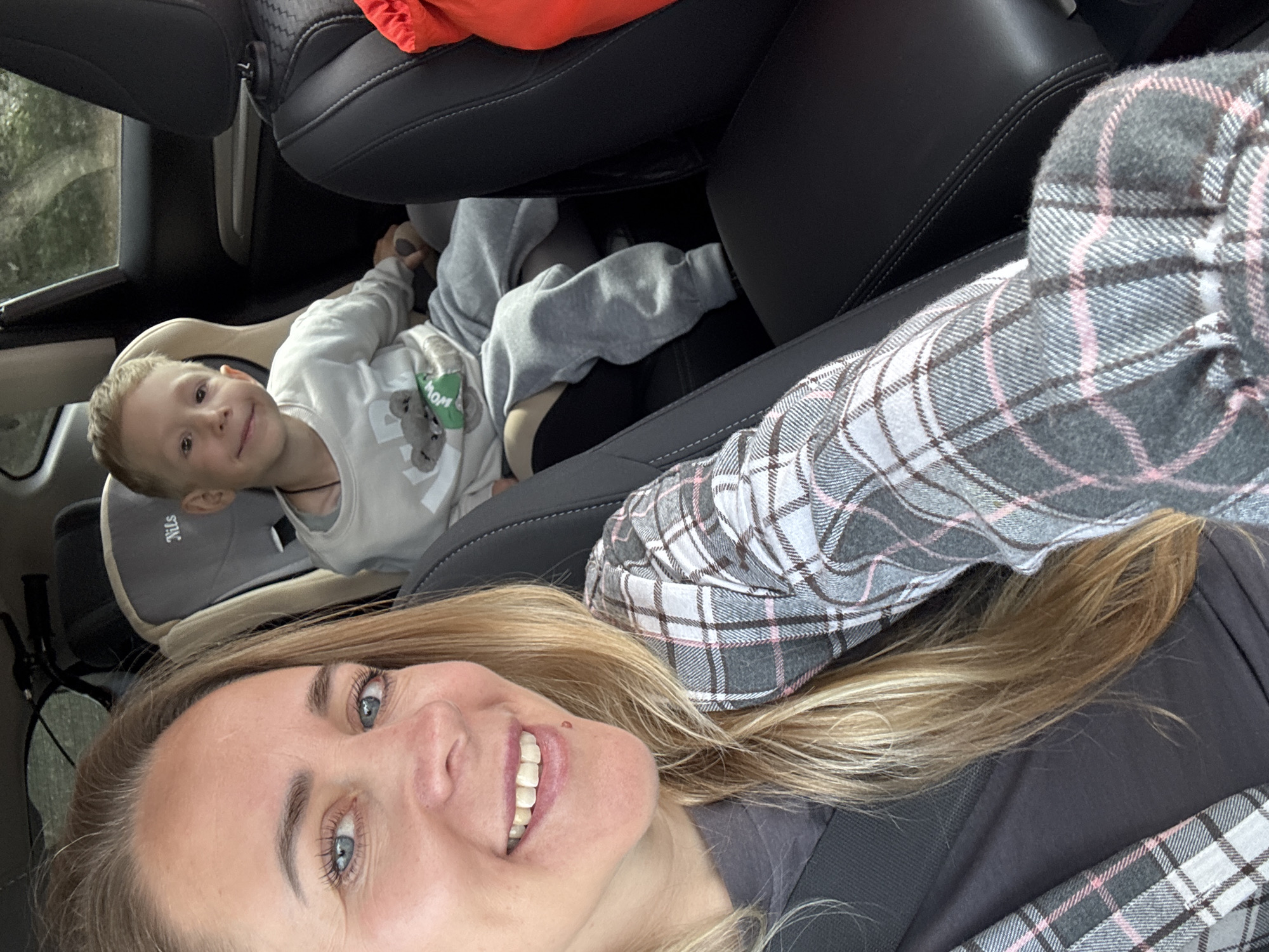 A woman is taking a selfie in a car. She is in the front passenger seat, and a young boy is in a car seat in the back. The woman has blonde hair and is smiling. She is wearing a plaid shirt. The boy is wearing a gray sweatshirt with a bear graphic. The car appears to be parked. The image was taken in Chaikovsky, Russia.