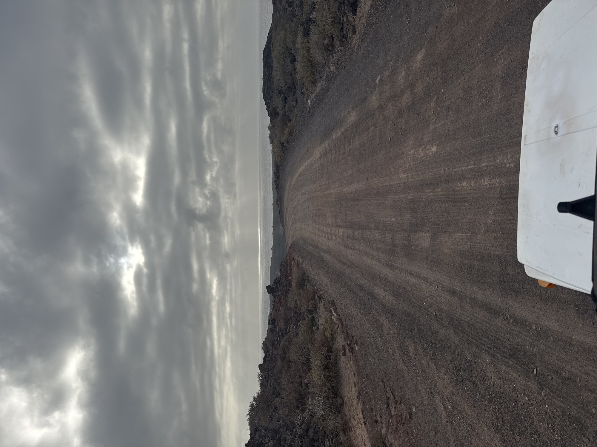 The image shows a dusty, unpaved road curving along a rocky coastline in the municipality of Tinajo, Spain. The sky is overcast with striated clouds. To the right is the side of a white vehicle. Sparse vegetation clings to the rocky terrain along the road. The sea is visible as a narrow band between the sky and the land.