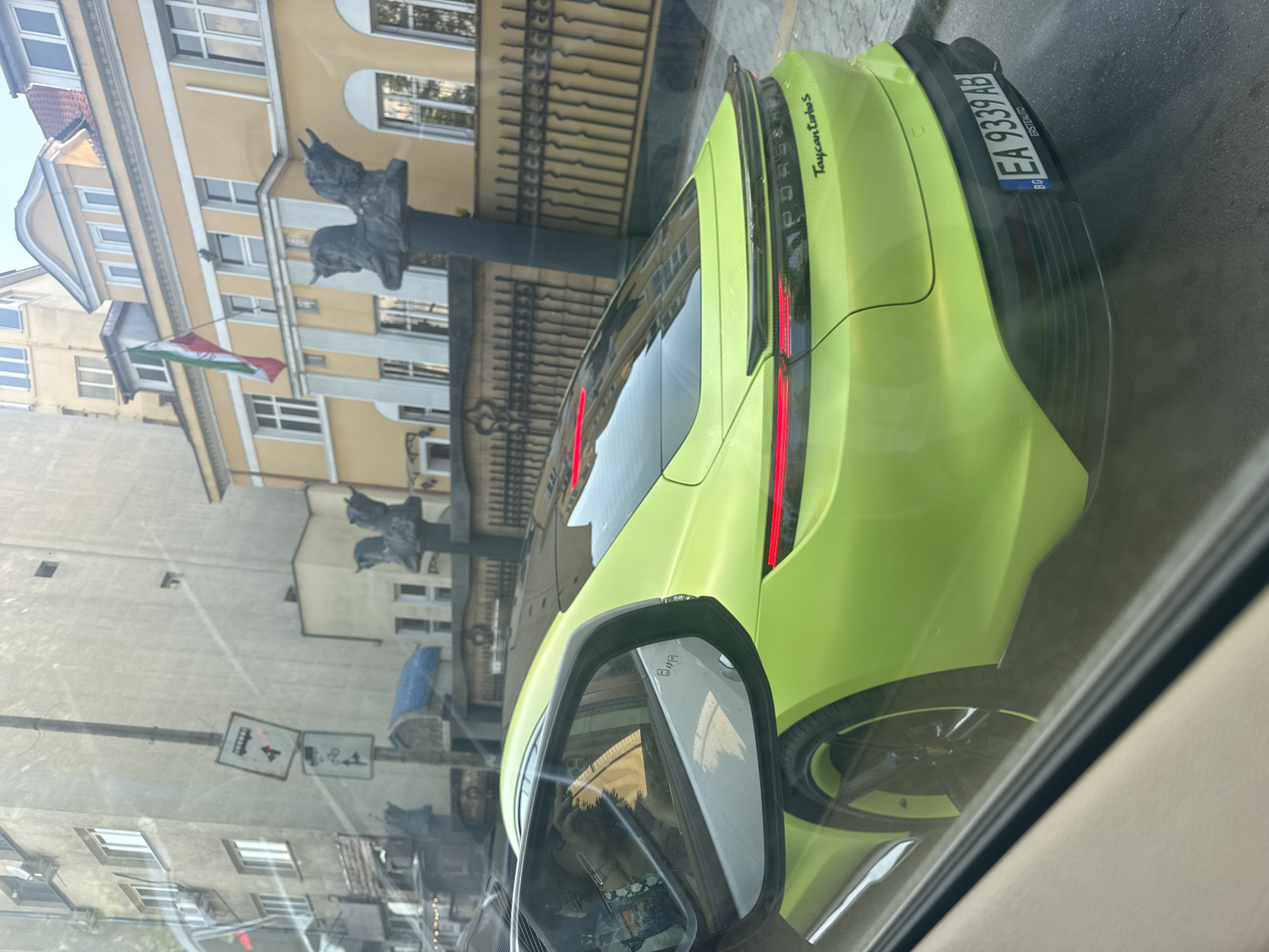 The image captures a bright green Porsche Taycan Turbo S driving through the city of Sofia, Bulgaria. The car is in motion, as indicated by the motion blur on the wheels and the reflection in the side mirror. The license plate is visible and reads "EA 9339 AB". In the background, an ornate yellow building with a red, white, and green flag flying from its facade stands out. The building is adorned with decorative sculptures. Traffic signs are also visible, including one indicating a tram crossing and another related to cycling and pedestrian pathways. The scene appears to be a typical urban street with buildings and infrastructure characteristic of a European city. The perspective suggests the photo was taken from inside another vehicle.