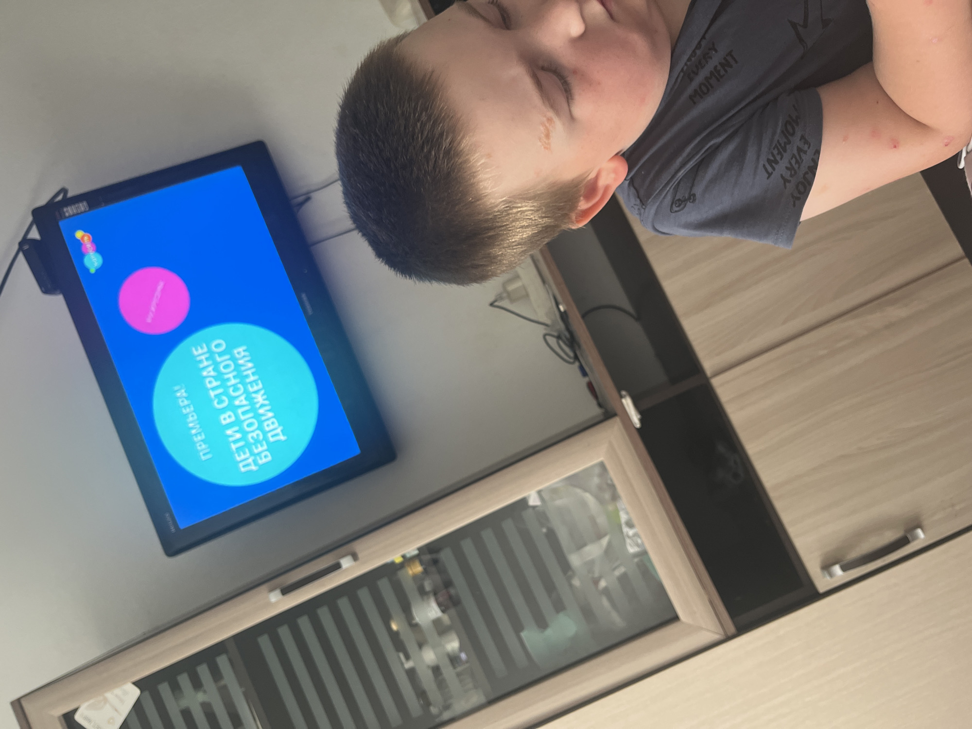 An eye-level medium shot captures a young boy from the shoulders up, sitting in front of a television in a room with light-colored wooden furniture. The boy, with light brown hair, is looking down and to his right, his eyes closed, and his skin appears slightly flushed with what might be a mild rash on his face and arms. The television screen displays a bright blue background with text in Russian that translates to "PREMIERE: Children in the country of safe movement." A pink circle is visible on the screen, as are some colorful graphics at the top. To the left of the television, a tall, light-wood cabinet with glass doors containing various items is visible. The background consists of a plain wall and the furniture. There is no context to suggest the location is Chelyabinsk, Russia.