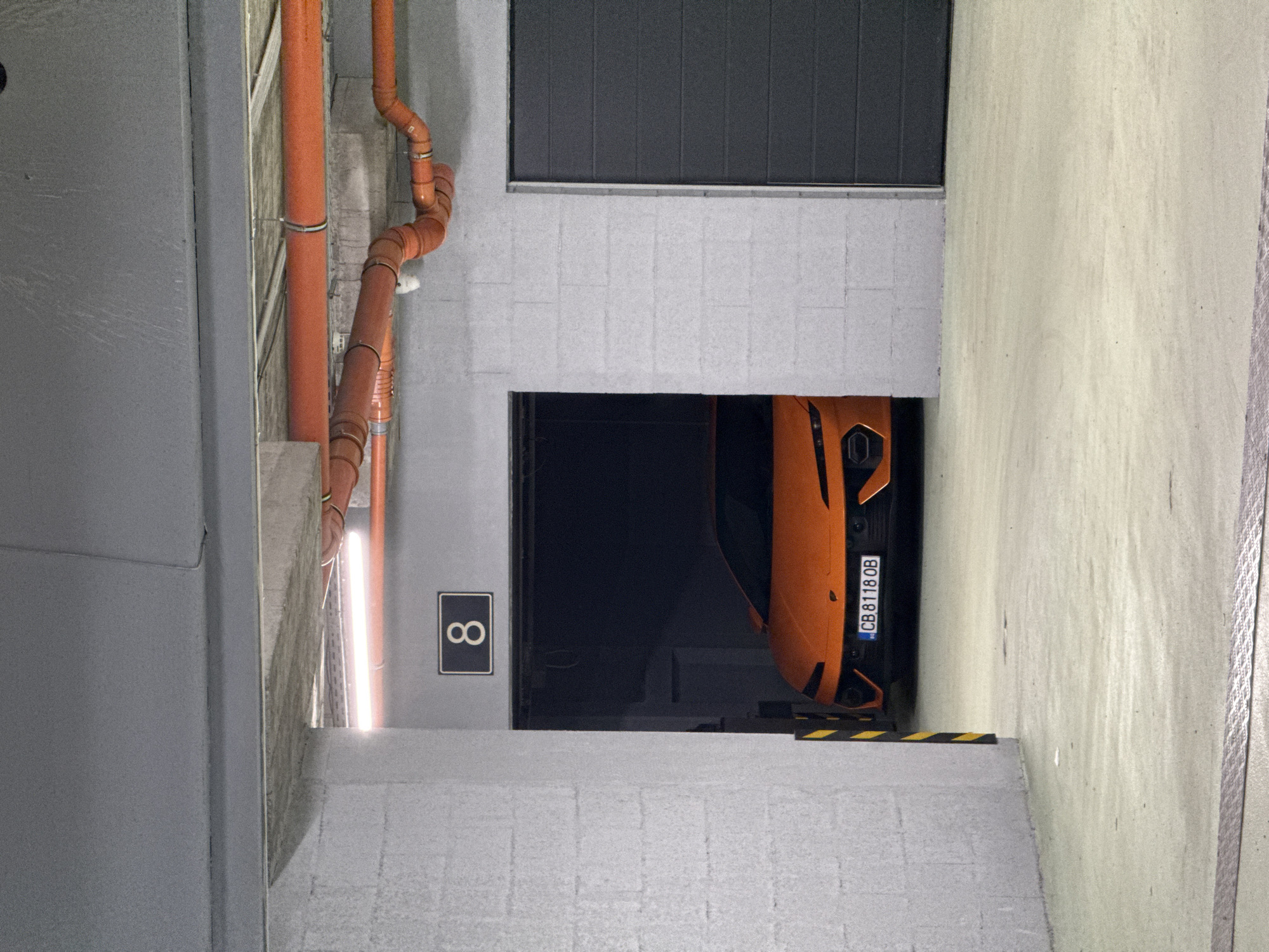 The image depicts a parking garage in Sofia, Bulgaria. A bright orange sports car is parked in parking space number 8. The car's license plate reads "CB 8118 OB". Orange pipes run along the left wall of the parking space.