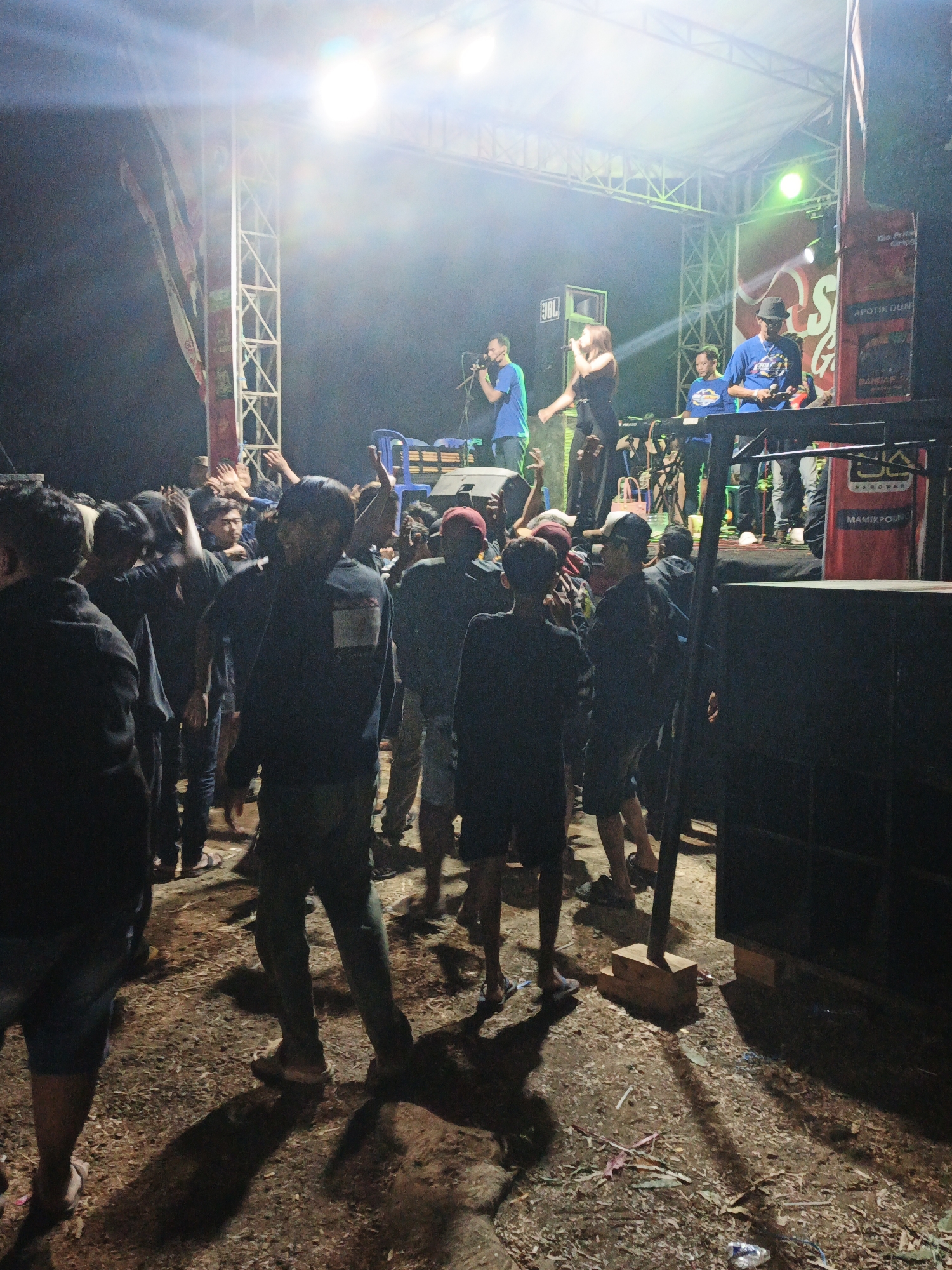 A nighttime outdoor concert is taking place on a makeshift stage in the city of Kepel, Indonesia. The stage is constructed with metal scaffolding and adorned with banners. Several people are on the stage, including musicians performing and what appears to be a DJ. The audience, a crowd of young men, is gathered in front of the stage, some with their hands raised in the air, suggesting they are enjoying the performance. The ground is covered in dirt, debris, and some scattered confetti. The lighting from the stage is bright and casts long shadows. The overall atmosphere appears to be lively and energetic.