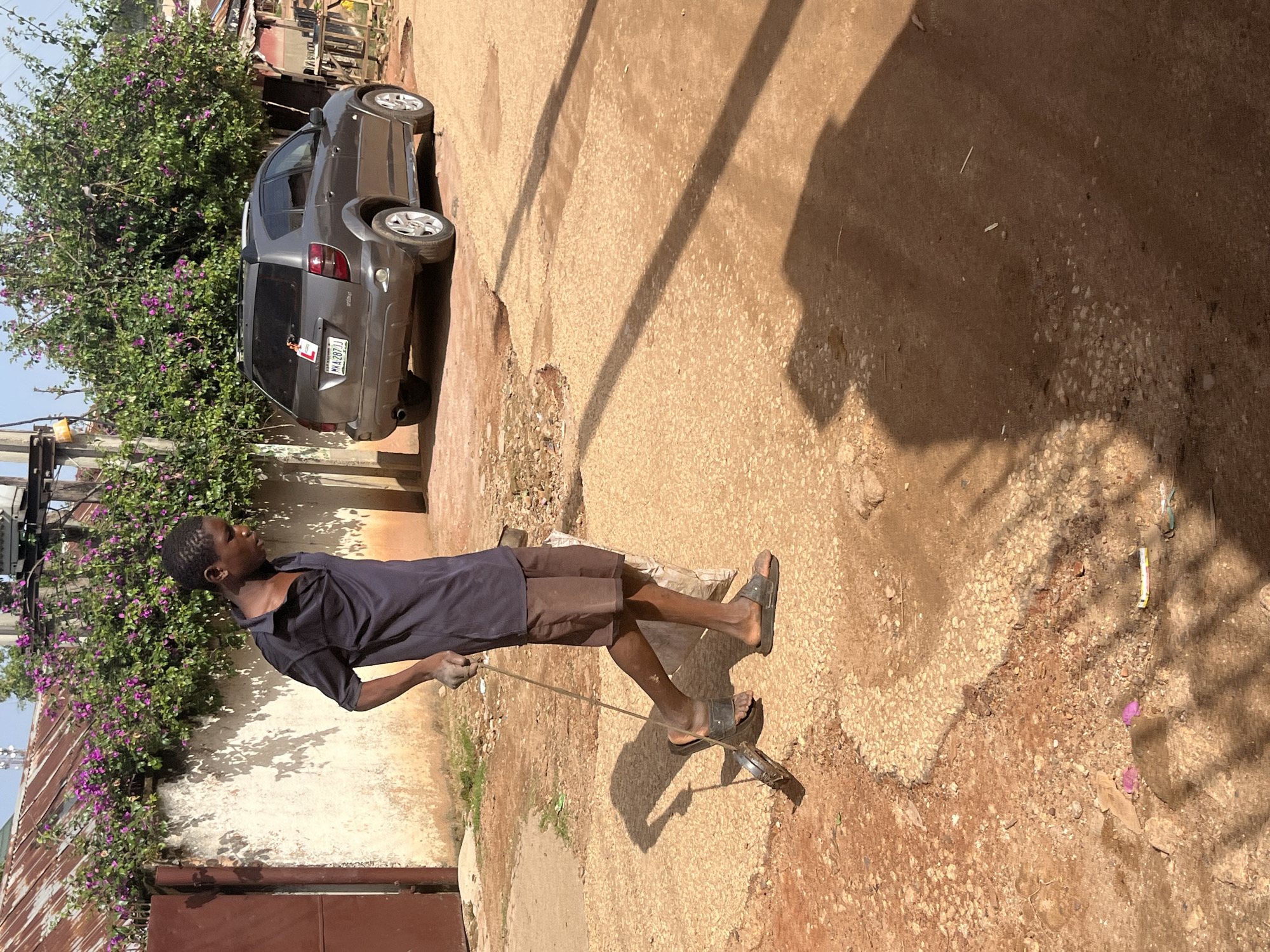 A young Black male, wearing a navy blue polo shirt and brown shorts, walks to the left across a dirt road in Jos, Nigeria. He is holding a metal rod with a circular metal object attached to the end, which he is using to sweep the ground. His feet are bare, and he is wearing dark colored sandals. In the background, a grey SUV is parked on the right side of the road. A white wall with vibrant purple flowers growing along its top runs along the left side of the road, with a large brown metal gate visible on the far left. The road is unpaved and dusty, with patches of gravel and some debris. The sun is shining brightly, casting long shadows across the ground.
