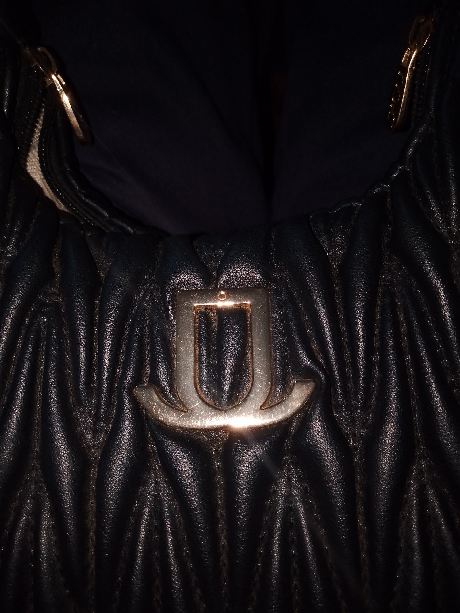 The image is a close-up, low-angle, dimly lit, indoor shot of a black, quilted leather handbag. The handbag features a prominent gold-toned "U" shaped emblem in the center, with a textured finish. To the left and right of the emblem, parts of a zipper and a gold-toned buckle are visible. The handbag is likely situated in a domestic setting in Tacarigua, Venezuela, given the context of the request, though no external environmental cues are present in the image itself to confirm this location. The focus is entirely on the design and material of the handbag.