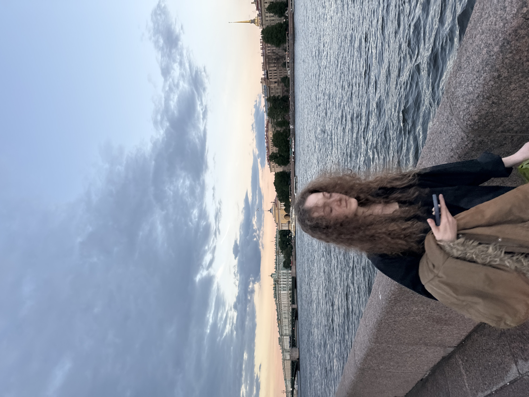 A young woman with long, curly brown hair stands on a granite embankment next to a body of water. She is wearing a black top and holding a brown jacket with a fur-lined hood in her left arm. Her right hand is holding a smartphone. In the background, across the water, is the city of Saint Petersburg, Russia. The cityscape features a long, multi-story building with classical architecture, a bridge, and several other buildings, including one with a golden spire. The sky above is filled with clouds and hints of a sunset with soft orange and pink hues. The water is dark and slightly rippled, reflecting the sky and buildings. The lighting suggests it is late afternoon or early evening.