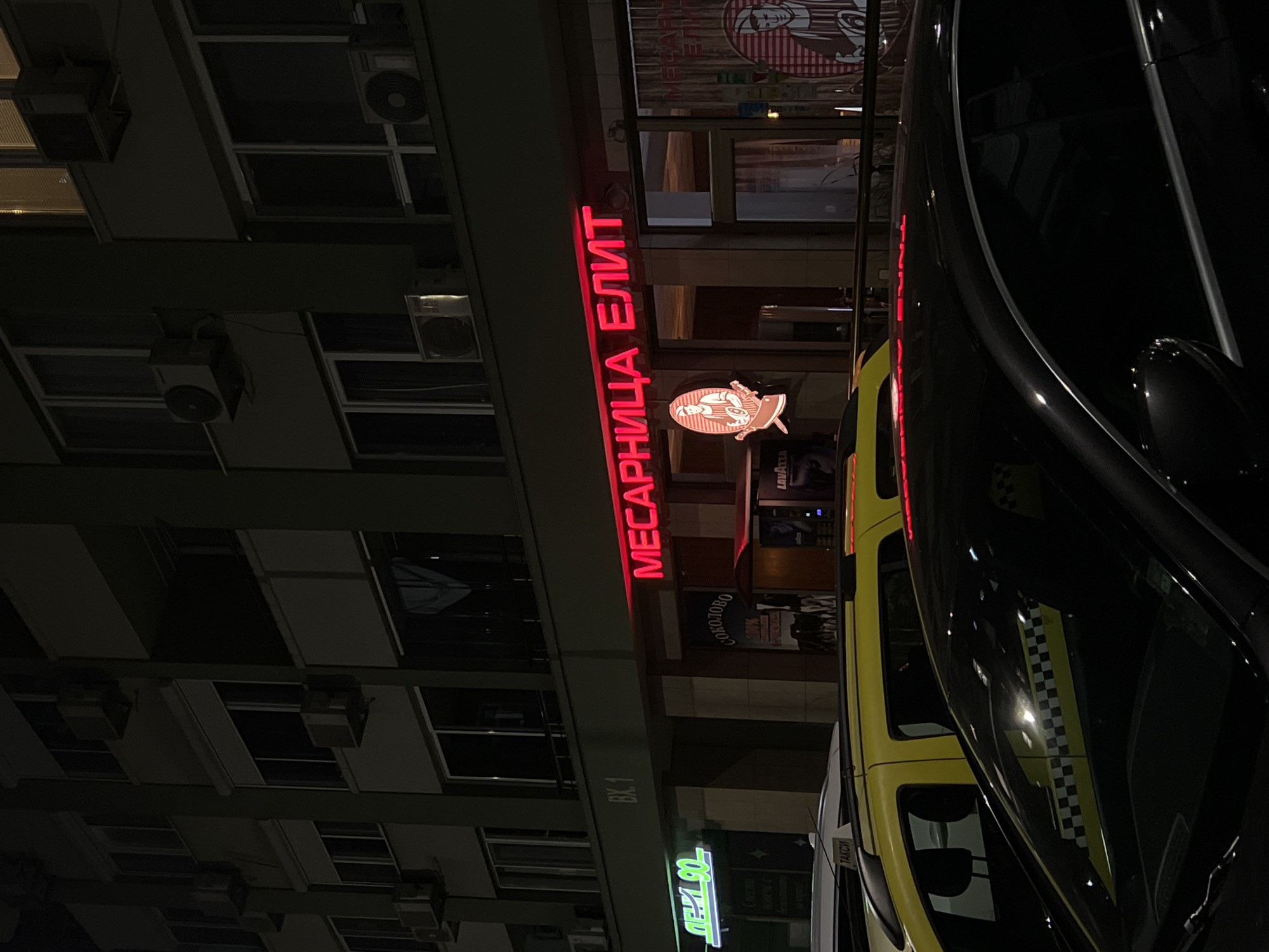 A nighttime street scene in Burgas, Bulgaria, features a multi-story residential building with air conditioning units visible on its facade. In front of the building is a lit sign in Cyrillic letters, "МЕСАРНИЦА ЕЛИТ" ("Butcher Elite"), above a logo depicting a butcher. Adjacent to the building, another sign with Cyrillic letters, "ЛЕВ 90", is seen. A black car and a yellow taxi are parked on the street, partially obstructing the view of the storefronts behind them. The environment is dark, with artificial lighting from the signs and reflections on the vehicles.
