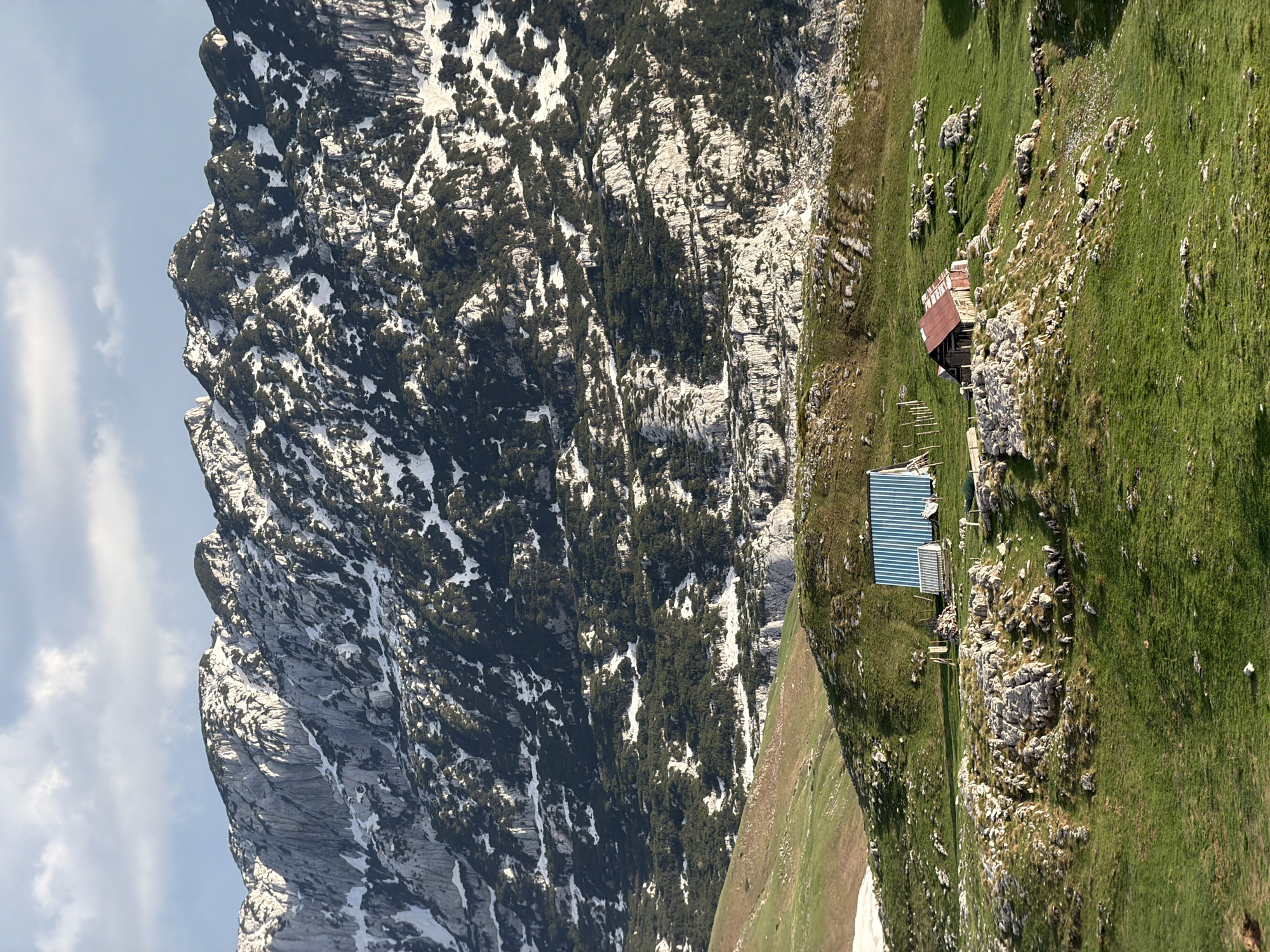 The image depicts a rugged mountain landscape, likely in the vicinity of Komarnica, Montenegro, though specific location is difficult to ascertain without more context. The left portion of the image is dominated by a steep, rocky mountain face with patches of snow and sparse vegetation clinging to its sides. To the right, the terrain transitions to a grassy slope interspersed with rocks and two small buildings. One building has a rusty red roof, and the other has a striped blue roof. The sky is visible in the upper left corner.