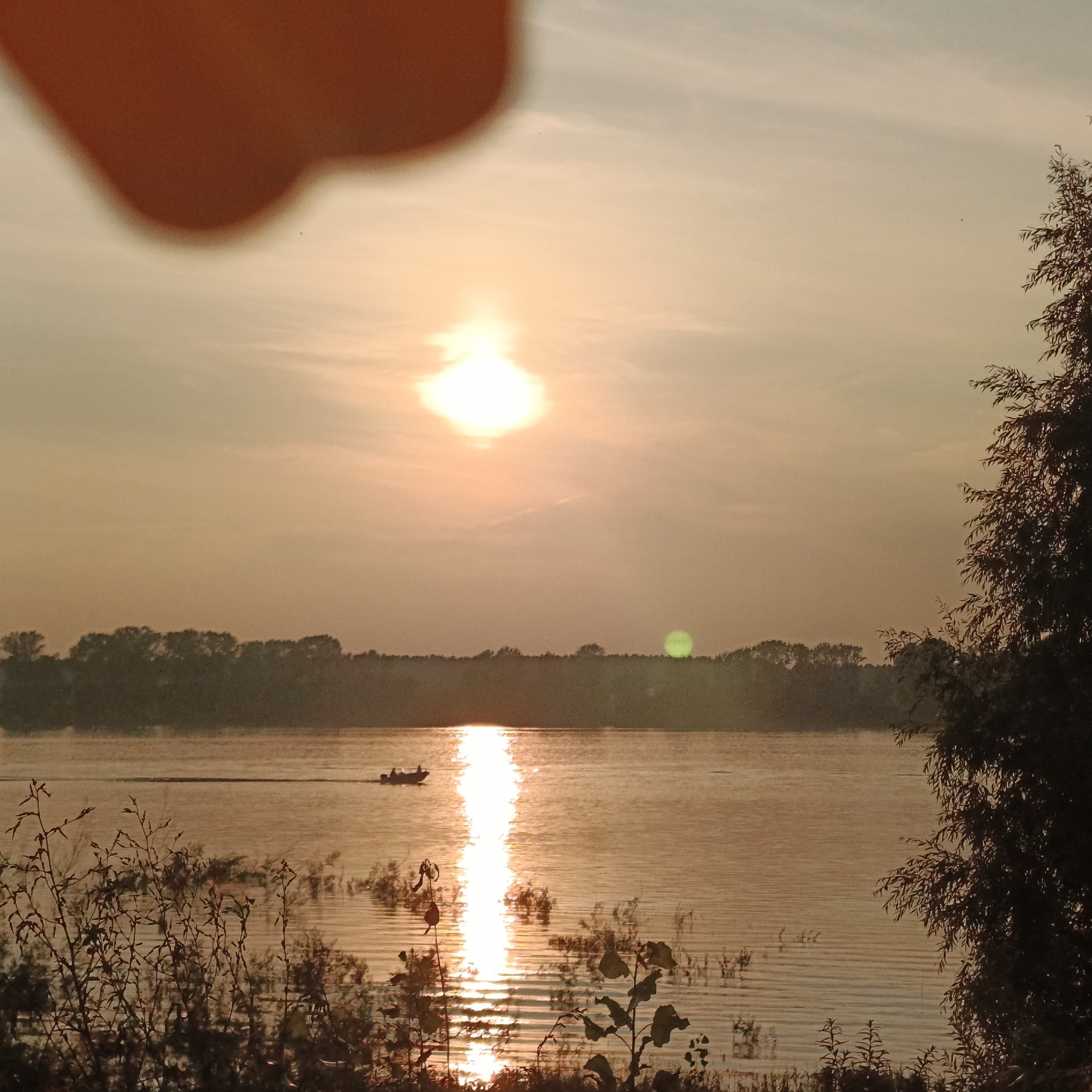 A sunset scene unfolds over a wide body of water, likely the Kama River, near Chaikovsky, Russia. The sun, partially obscured by a hazy atmosphere and possibly a foreground element like a finger, hangs in the sky, casting a warm, orange glow that reflects brightly on the water's surface. A thin, dark line of trees forms a distant horizon. In the middle ground, a small motorboat with at least two occupants is visible, moving across the water, creating a wake that interacts with the sun's reflection. The foreground is partially filled with dark, silhouetted vegetation, some of which appears to be on the bank of the river. A large, blurry orange shape dominates the upper left quadrant of the image, suggesting a hand or object is close to the camera lens, partially obscuring the sky.