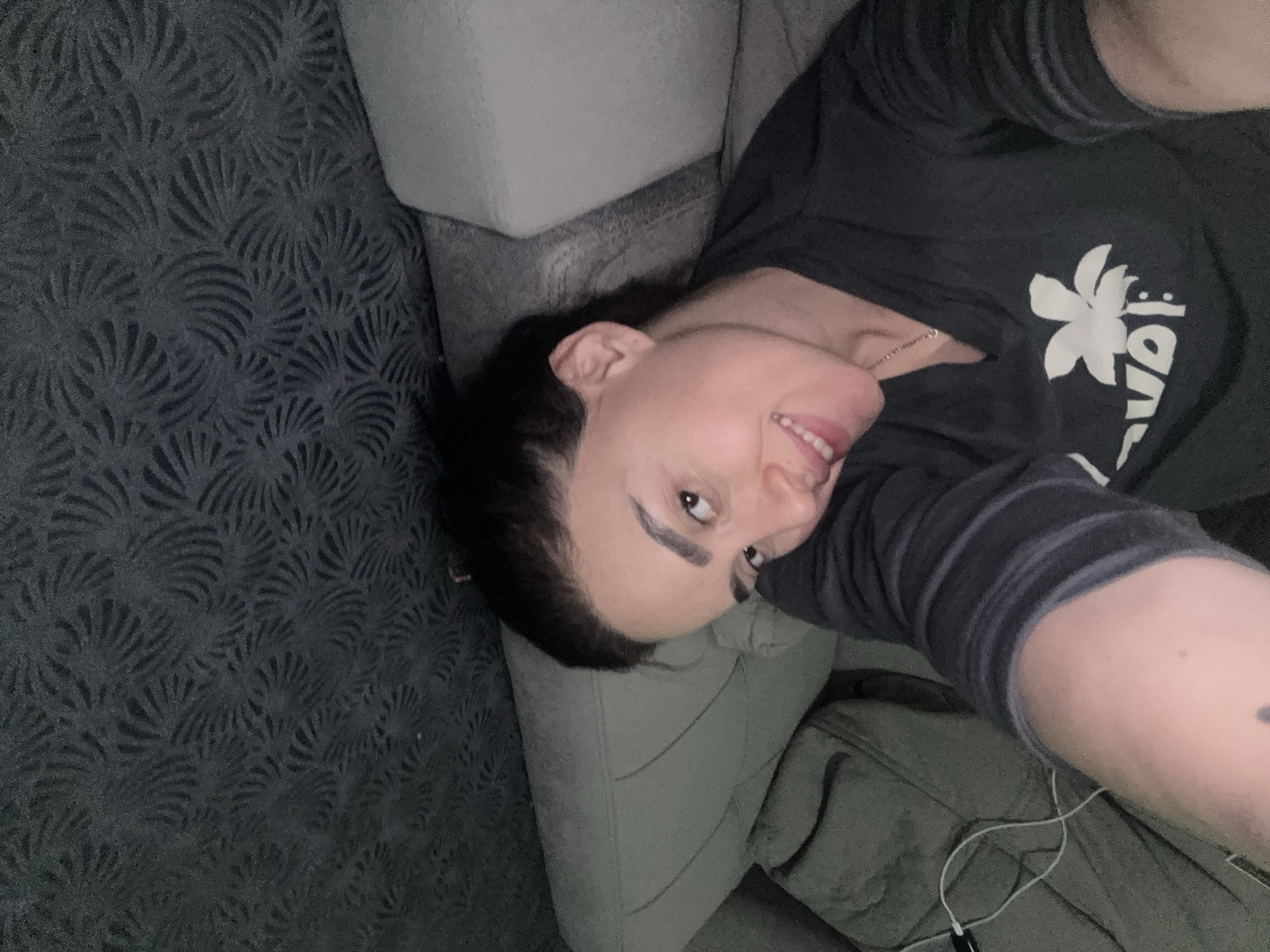 A person lies on a grey patterned couch. The person wears a dark-colored shirt with a white flower graphic and a striped sleeve. An earbud wire is visible near their arm. The location is impossible to determine from the image.
