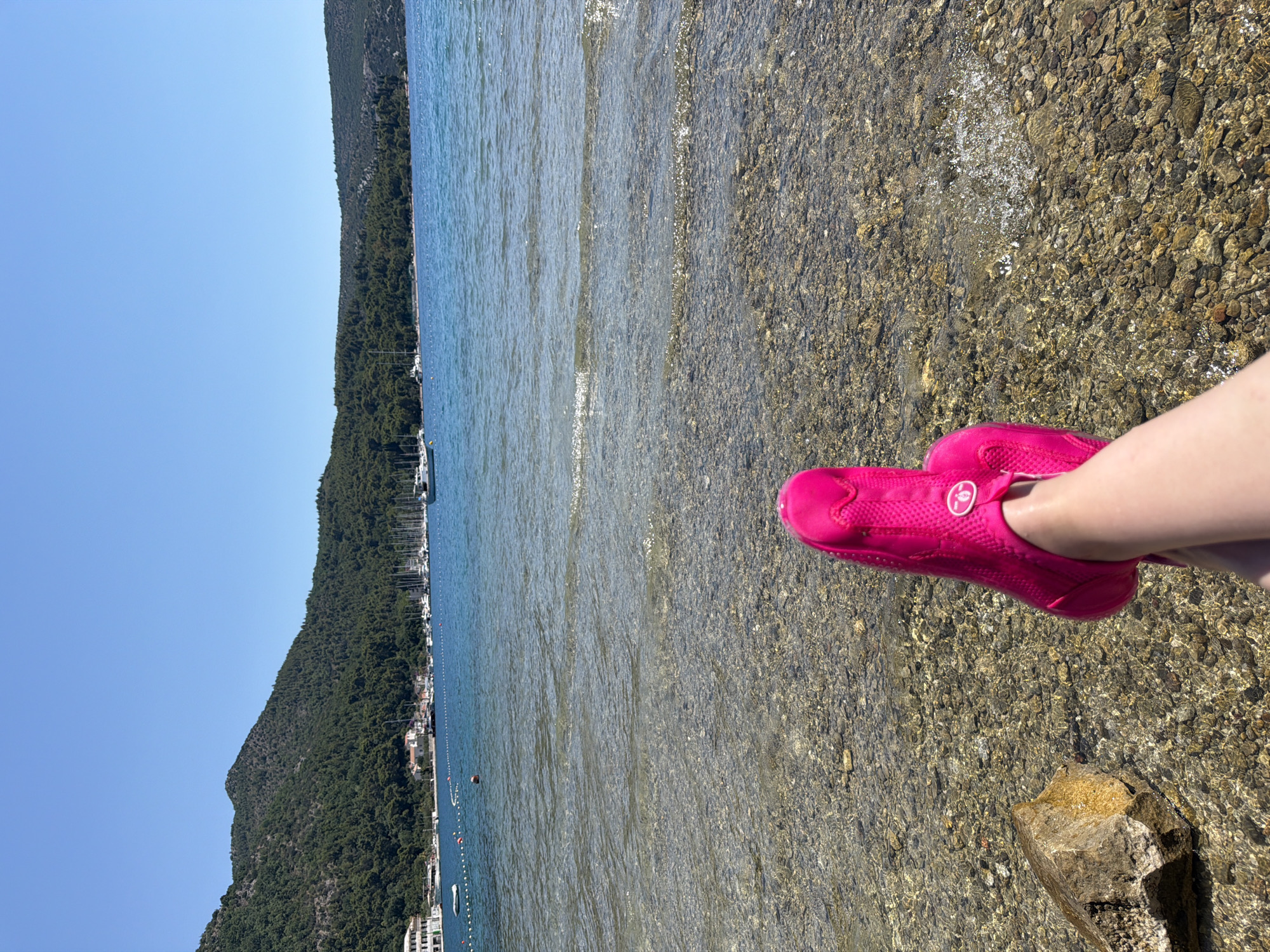 A person's feet, wearing bright pink water shoes, are visible in the foreground, resting on a rocky beach at the edge of clear, shallow water. The water is calm with gentle ripples, and the seabed is composed of small, multi-colored stones. Beyond the shoreline, a lush, green, tree-covered mountain slopes down towards the sea. A line of buildings and boats is nestled along the coast at the base of the mountain, suggesting a small town or village. The sea stretches out to the horizon, meeting a clear, bright blue sky. The sun appears to be high in the sky, casting shadows and illuminating the scene with bright light, indicating it is likely midday on a sunny day. The overall impression is a peaceful and idyllic coastal scene, likely a vacation destination. No visible text is present in the image.
