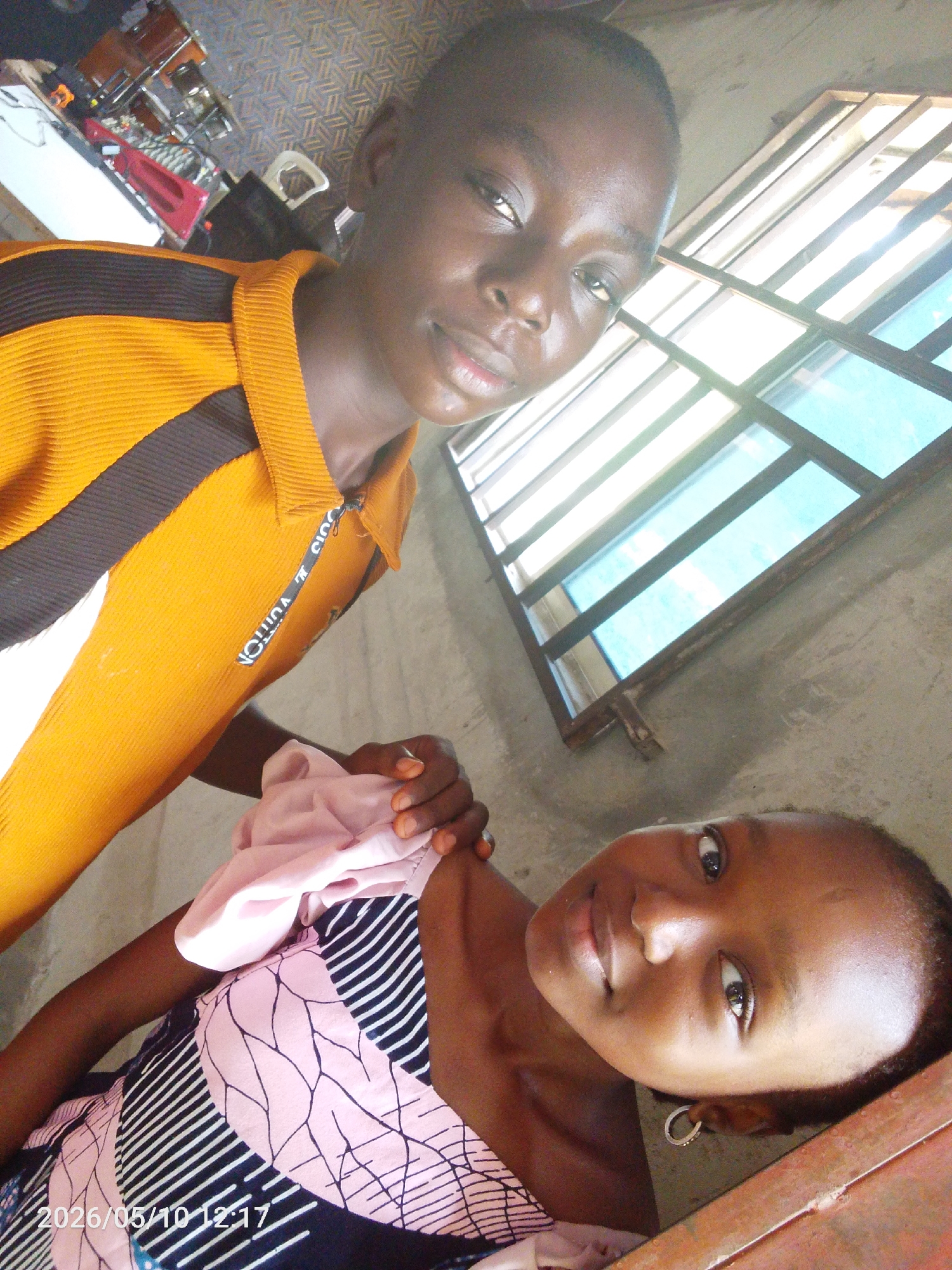 Two young individuals are captured indoors. The person on the left wears an orange and black shirt, while the person on the right is dressed in a pink and black patterned top. The environment appears to be indoors with a window providing natural light. A date stamp of "2026/05/10 12:17" is visible. The location in the city of Lokoja, Nigeria is not identifiable from the image.