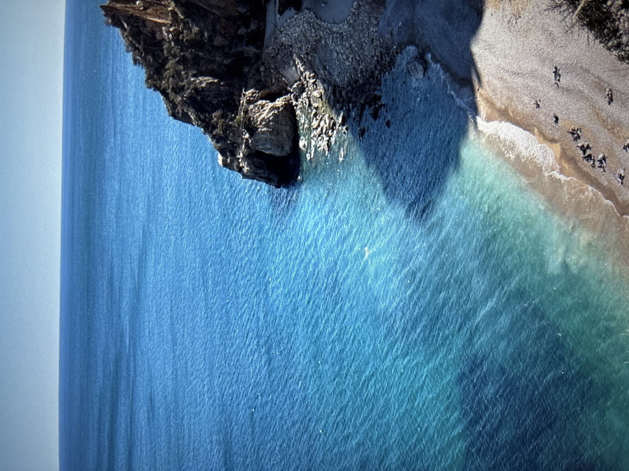 This is an aerial shot of a rocky coastline with clear blue water. A small, pebbly beach is visible in the foreground, with several scattered figures sunbathing. The water transitions from a bright turquoise near the shore to a deep blue further out. A rugged, dark cliff face juts out into the sea on the right side of the image, featuring a cave-like opening. The sky above is a pale, cloudless blue. This image is not of Sofia, Bulgaria.