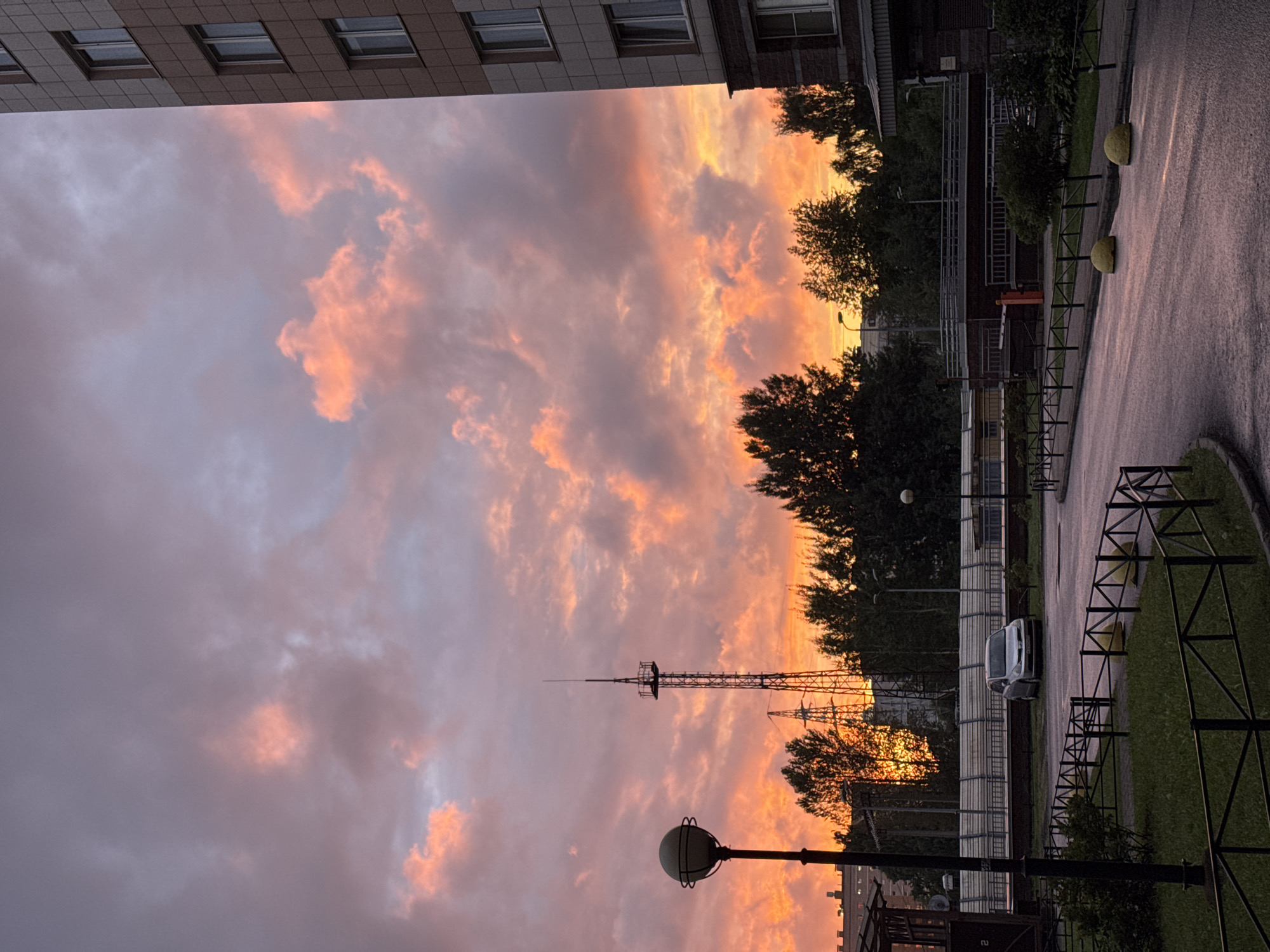 The image depicts a dramatic sunset over a residential area in an unknown city in Russia. The sky is filled with vibrant colors, ranging from deep orange and yellow near the horizon to soft pinks and purples higher up, with dark, textured clouds. In the foreground, a wet asphalt road curves to the right, bordered by a black metal fence and patches of green grass. A silver car is parked on the side of the road. To the left, a lamppost with a spherical head stands prominently. Behind the car and fence, there is a line of trees and a tall radio tower. To the far right, a portion of a multi-story apartment building with light-colored facade and visible windows is visible. The overall scene is one of an urban environment at dusk, with a striking natural phenomenon dominating the sky.