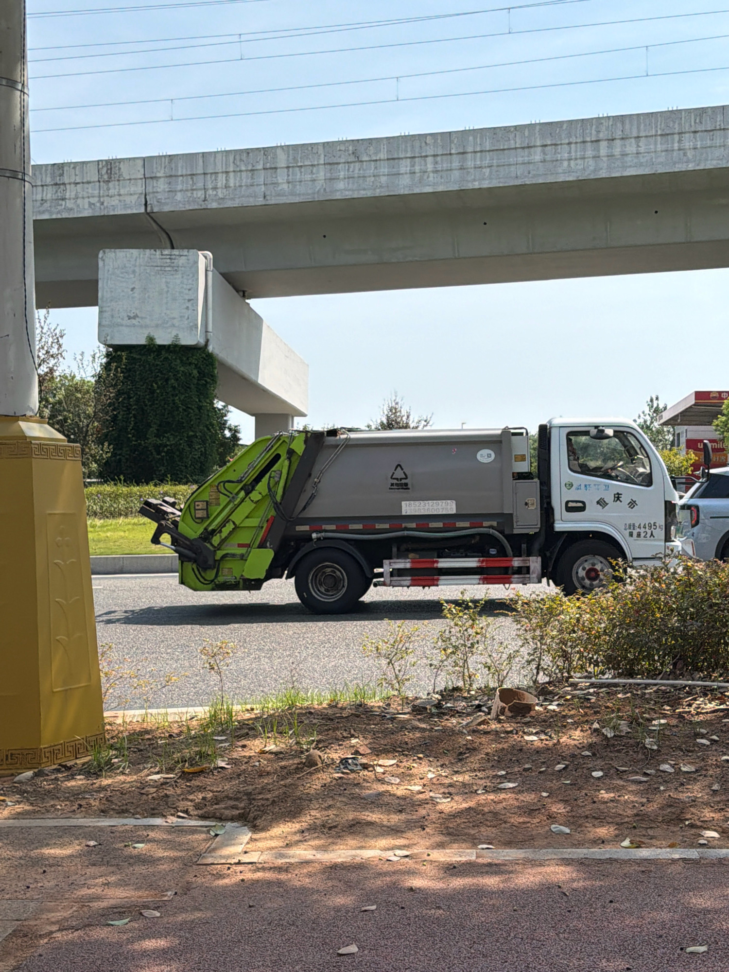 The image depicts a white and gray garbage truck parked on an asphalt road, partially obscured by green foliage and a yellow pillar on the left. The truck has a green mechanical arm at the rear, used for lifting and compacting waste. The background reveals a concrete overpass with power lines above it, suggesting an urban or semi-urban setting. Text is visible on the side of the truck, including Chinese characters and numbers, possibly indicating the vehicle's registration or company information. A silver car is parked behind the garbage truck on the right. The ground in the foreground is a mixture of dirt, grass, and fallen leaves, with a sidewalk visible at the bottom edge. The sky is clear and blue, indicating daylight. Based on the presence of the overpass, power lines, and the style of the truck, the scene is likely in Jiangjin, China. There are no people clearly visible in the image.