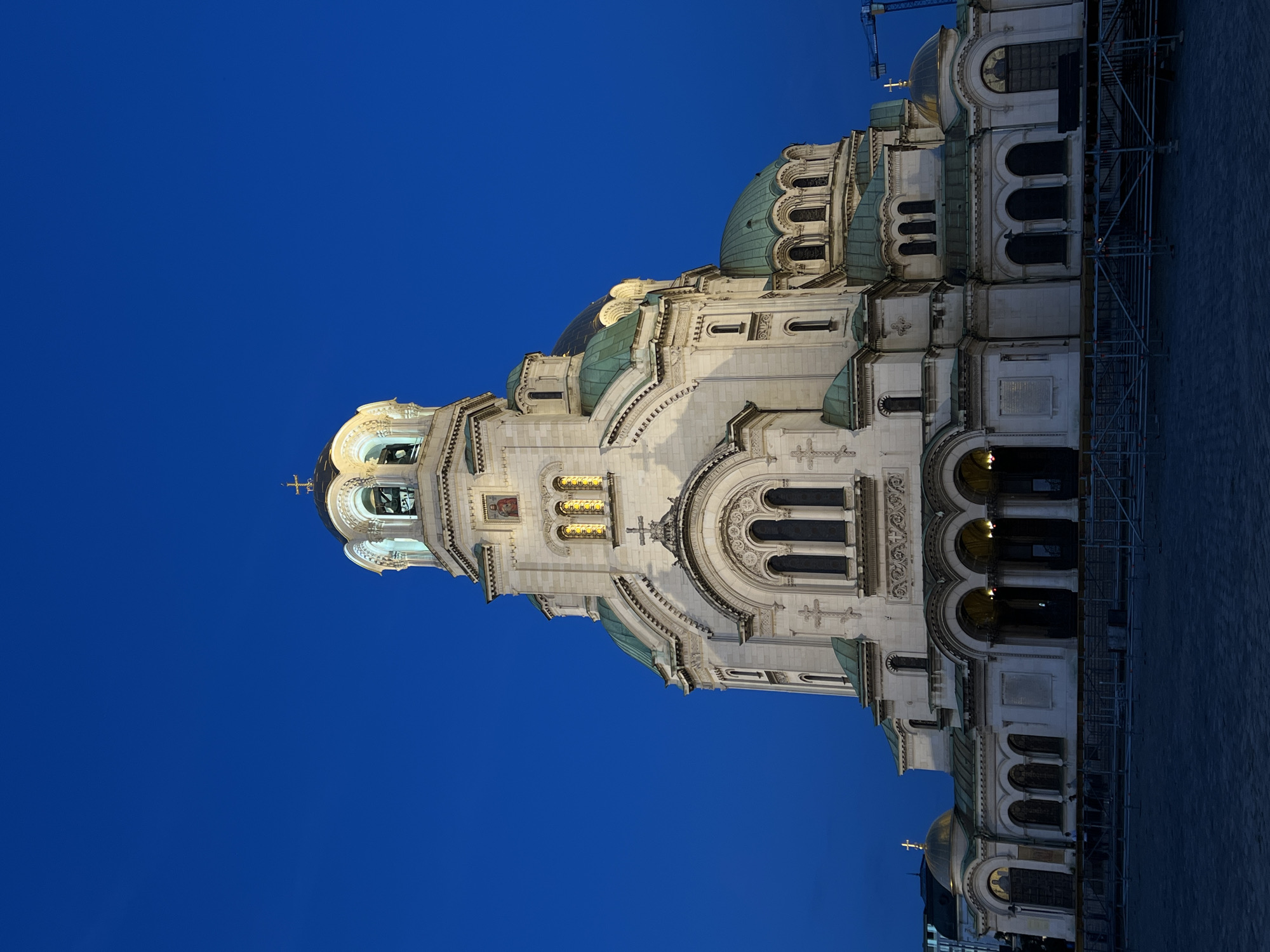 The image shows the Alexander Nevsky Cathedral in Sofia, Bulgaria, at dusk. The cathedral is a large, white, Orthodox church with multiple domes, a prominent central tower with a bell, and a golden dome with a cross. The sky behind the cathedral is a deep blue. The foreground shows a paved plaza with scaffolding and some temporary barriers. The lighting suggests the image was taken in the late evening or early morning. No people are clearly visible, though there are some faint figures in the lower left section of the image. The overall scene depicts a prominent landmark in Sofia, illuminated against a dark sky.