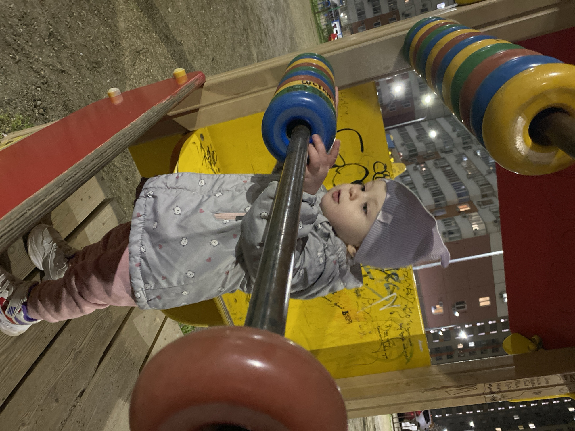 A young child, wearing a grey jacket with small heart and animal designs, pink pants, white sneakers, and a lavender hat, stands on what appears to be a wooden structure in a public park or playground. The child is grasping a vertical metal bar adorned with large, colorful plastic rings. The structure also includes a red slide and a yellow panel featuring graffiti or markings. In the background, there is a multi-story residential building with multiple lighted windows. The environment suggests a nighttime setting, potentially in an urban residential area. The city and specific region are unknown. The child's action is interactive, engaging with playground equipment.