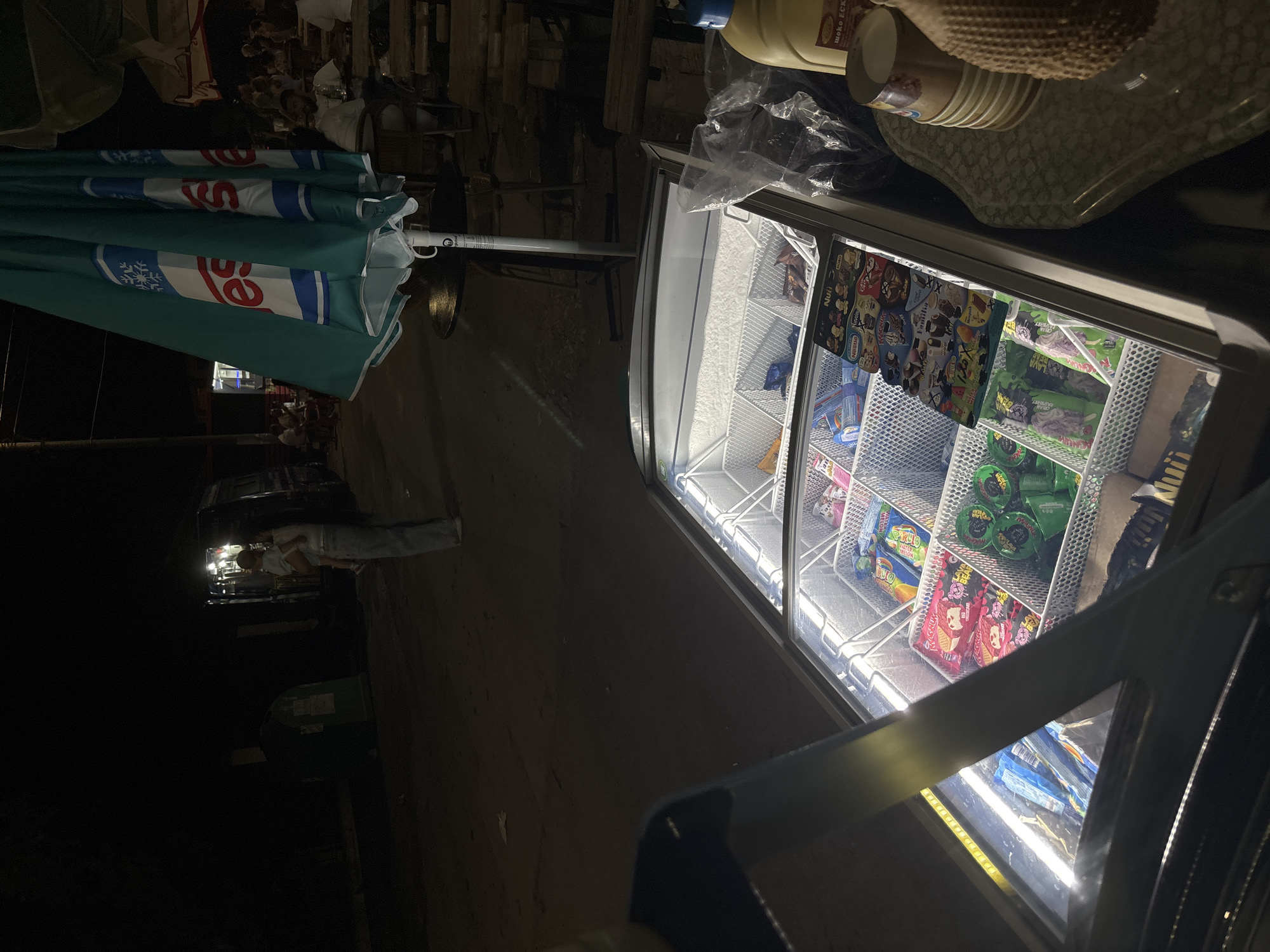 This image depicts an outdoor night scene in Kostievo, Bulgaria. In the immediate foreground, a white, glass-top freezer is illuminated internally, displaying various packaged ice cream products and popsicles arranged in white wire baskets. Visible brands include Nestle, Nui, and MBO, along with a laminated menu displaying additional ice cream options. To the right of the freezer, on an adjacent surface, are a yellow plastic bottle, a stack of small brown paper or plastic cups, and a textured, woven object.
A large, partially furled teal outdoor umbrella, branded with "ESKIMO" and a snowflake logo in white and blue, stands centrally on a pole. The ground in the foreground is dark and unpaved.
In the midground, towards the left, a woman with long dark hair, wearing light-colored jeans and a top, is holding a small child dressed in light attire. They are positioned near a dark van or utility vehicle with its interior lights on. To the right of the umbrella, an outdoor seating area is visible, with several individuals seated at tables, including a bearded man in a white t-shirt. Stacks of wooden pallets are present on the far right. The background beyond these elements is obscured by darkness, with a faint outline of a green dumpster visible in the far left. Artificial lighting sources illuminate the freezer, the vehicle, and the immediate seating area.