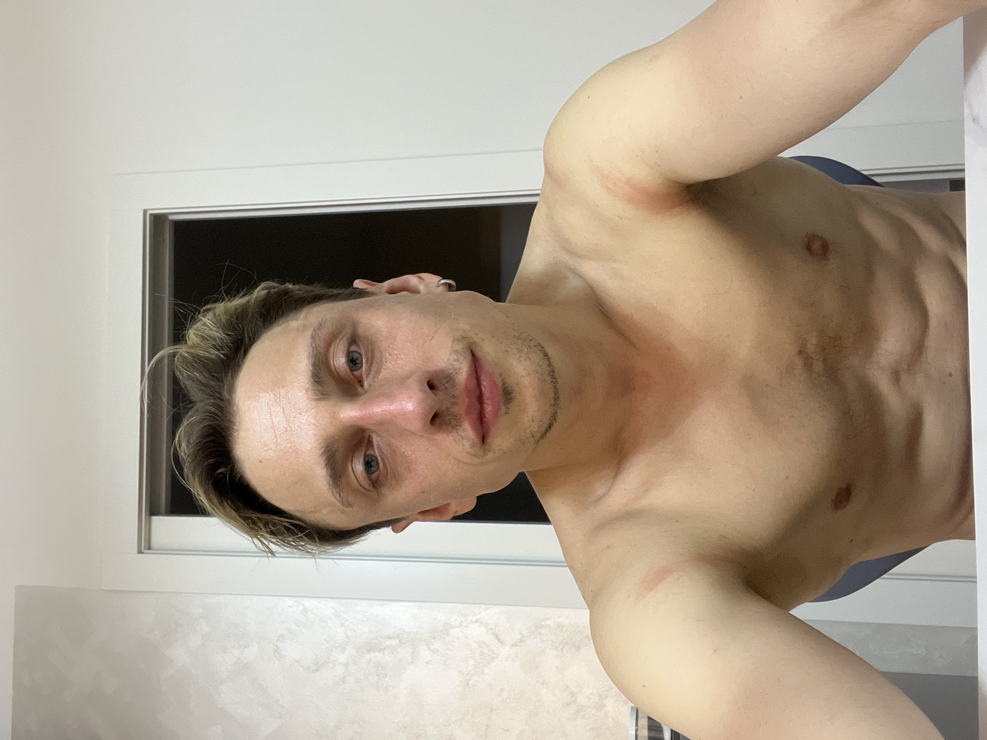 A shirtless man with light skin, blonde hair, and blue eyes is looking directly at the camera. He has a light mustache and stubble. There are red marks on his upper arms and shoulders. He is wearing a silver hoop earring in his left ear. The background is a plain white wall with a door frame. The scene appears to be indoors, possibly in a bathroom or bedroom. There are no identifiable landmarks or clues that would place this image in Verona, Italy.