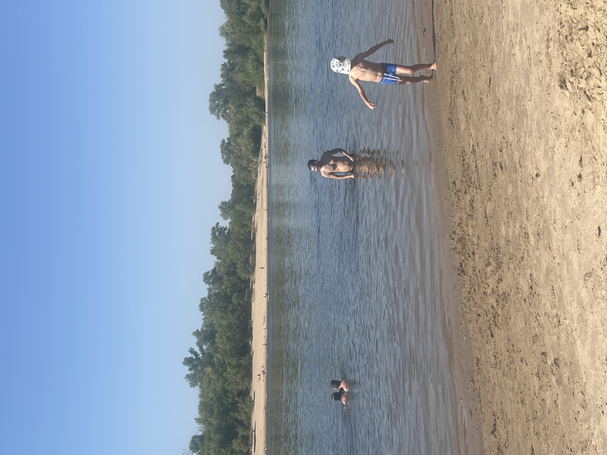 In Kilyakovka, Russia, on a clear, sunny day, people are enjoying the river. The foreground shows a sandy beach with visible ripples and texture. The water is calm and reflects the sky. Three individuals are visible in the water. Two people are partially submerged near the left side of the frame, with their backs to the camera. Further to the right, a man stands waist-deep in the water, facing towards the right. A young boy, wearing blue swim trunks and a patterned hat, is running towards the shore from the water, with his arms outstretched. In the background, a lush green treeline borders a sandy bank, where several other individuals are scattered, some sunbathing and others near the water. The overall scene depicts a peaceful, recreational day at the river.