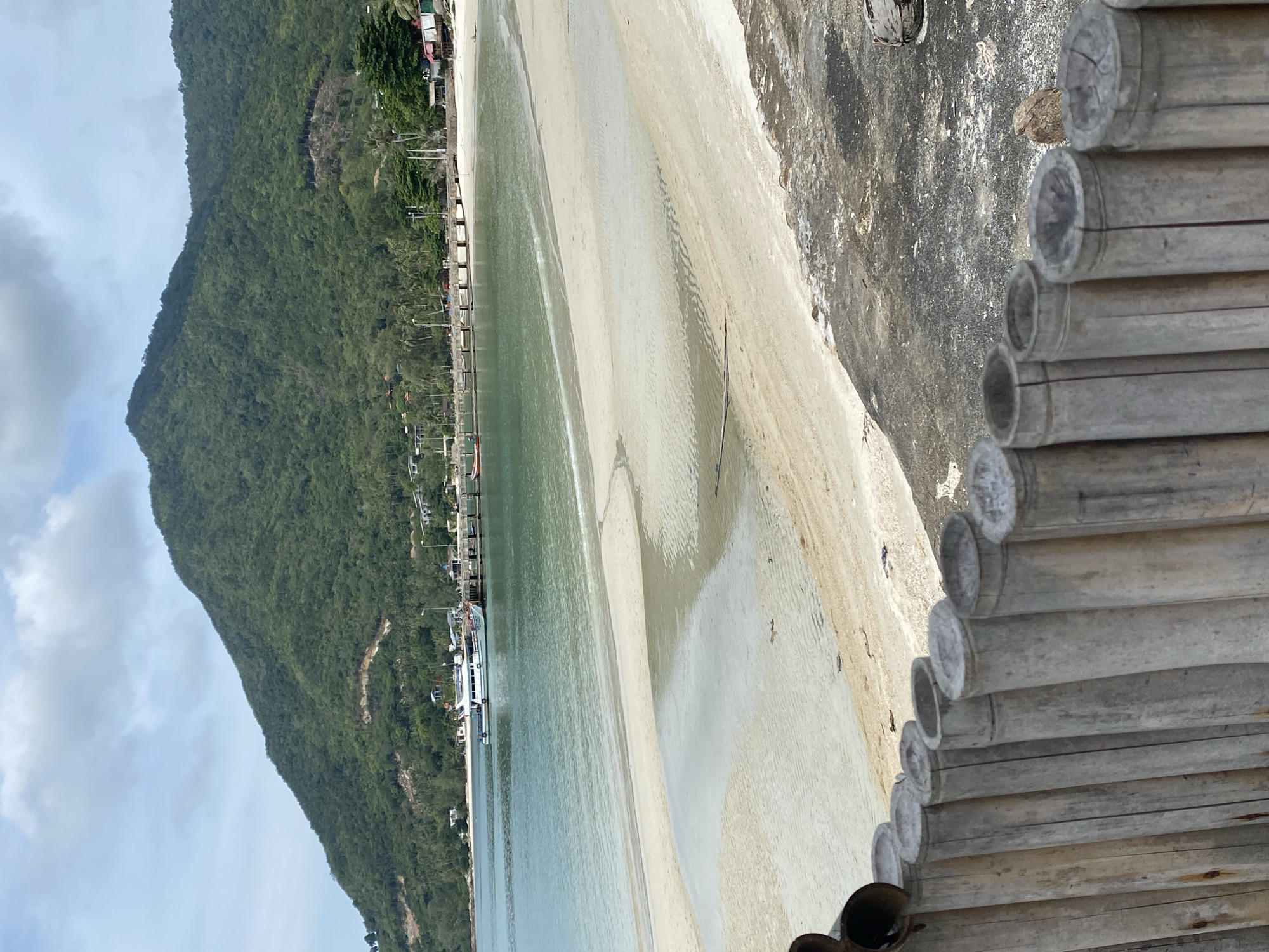 The image shows a view of a beach in Baan Chaloklum, Thailand. A mountain covered in lush greenery is on the left side of the frame. The beach is sandy and merges into the water, which is light turquoise. There is a long pier extending into the water with buildings and structures along its edge. Several boats are moored along the pier. In the foreground, there are bamboo poles arranged in a row, indicating a man-made structure like a fence or wall.