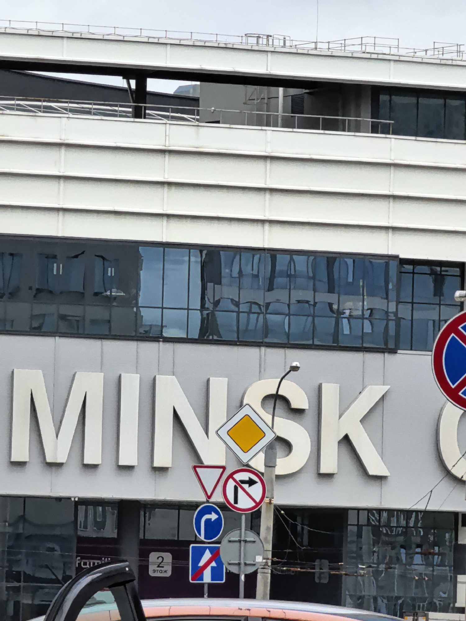 The image shows a close-up view of the facade of a modern building with large, reflective windows. The word "MINSK" is prominently displayed in large, white, three-dimensional letters on the building's exterior. Below the word "MINSK", there is a row of dark, reflective windows. Several traffic signs are visible in the foreground, including a diamond-shaped yield sign, a circular "no right turn" sign, and a rectangular sign with a blue circle and a white arrow pointing right, indicating a mandatory turn. A circular sign with a red border, a blue background, and a red diagonal cross is visible on the right side of the image. The building's architecture features horizontal white panels. The sky is overcast and appears to be a light grey. In the lower part of the image, a black car roof and part of a vehicle's windshield are visible. There are no people clearly visible in the frame.