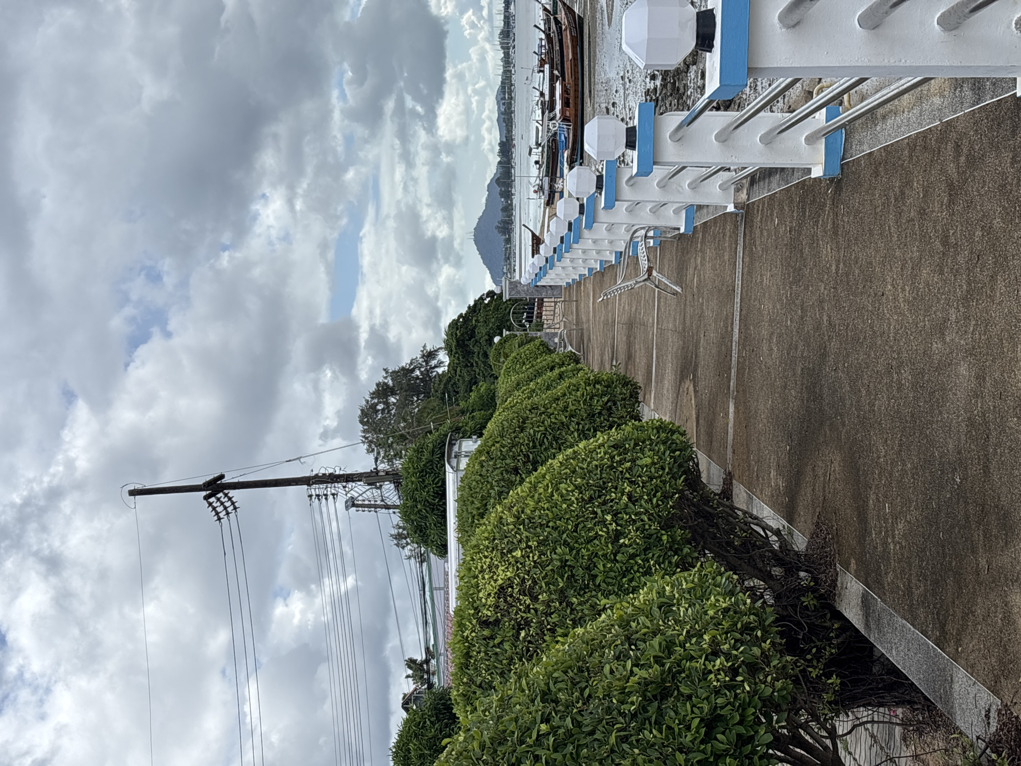 This is a medium shot of a waterfront promenade in Rawai, Thailand. The promenade is lined with manicured green bushes on the left and a white and blue railing with decorative lamps on the right. Beyond the railing, numerous longtail boats are moored in the shallow water, with a mountainous landscape visible in the distance under a cloudy sky. The ground of the promenade is made of textured concrete. There are two empty metal chairs on the promenade. Electrical poles and wires are visible in the background to the left.