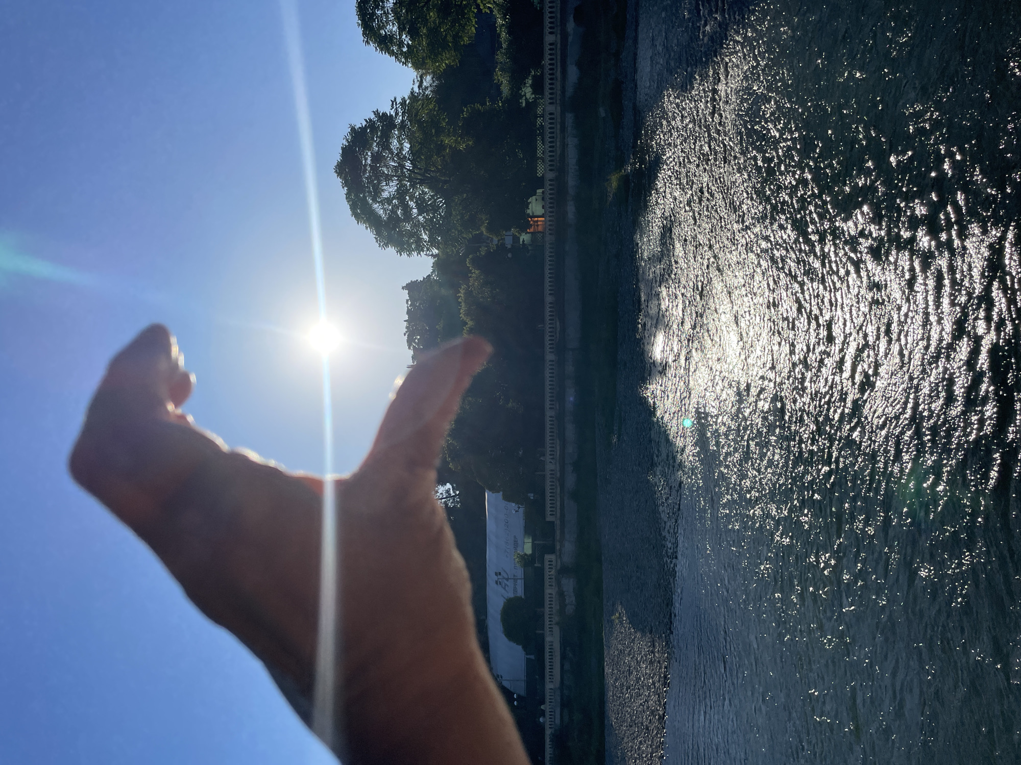 A low-angle, vertical shot captures a sunlit river scene in Sochi, Russia. A person's hand is silhouetted in the foreground, obscuring the upper left portion of the frame as it forms a crescent shape. The bright sun, partially blocked by the hand, creates a lens flare effect that streaks horizontally across the image. In the midground, a river flows, its surface shimmering with reflections of the sun. Along the opposite bank, a stone embankment is visible, topped by a low wall with decorative railings. Behind the wall, lush green trees and foliage form a dense backdrop. A white building with text on its side is partially visible amongst the trees. The sky is a clear, bright blue.