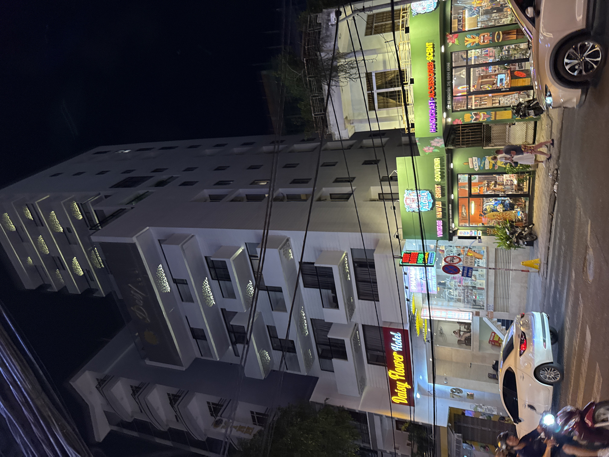 This is a night-time street-level photograph taken in Nha Trang, Vietnam, featuring the "Daisy Flower Hotel" and adjacent shops. The hotel is a tall, multi-story building with a lit sign that reads "Daisy Flower Hotel" in yellow, cursive lettering against a red background. The hotel's facade is white, with several balconies and windows illuminated from within. Below the hotel, a shop with a bright green facade displays the words "Aloha," "VINTAGE HAWAII SHIRT SOUVENIR," and "HANDICRAFT ACCESSORIES SCENT." This shop has large glass windows showcasing various merchandise, including clothing and decorative items.
In the foreground, a white sedan is parked and partially visible on the right side of the image. On the left, a person is riding a motorcycle with its headlight on. Another motorcycle is parked on the sidewalk in front of the green shop. Two individuals, a man and a woman, are walking on the sidewalk in front of the green shop. The street is paved, and there are streetlights and overhead power lines visible, contributing to the urban night-time atmosphere. The overall scene depicts a busy commercial street in Nha Trang at night.
