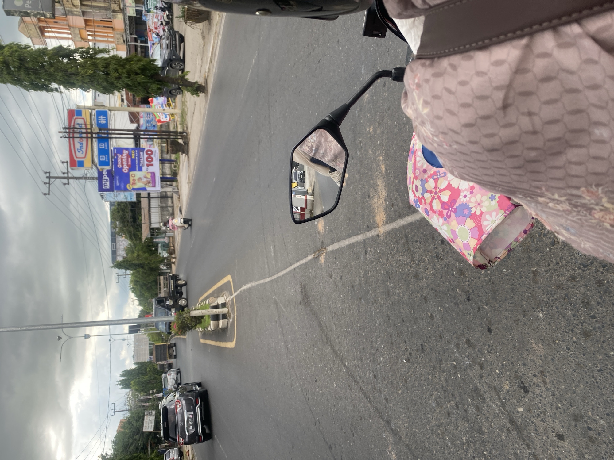 The image captures a street scene in Sungai Paring, Indonesia, under an overcast sky. The perspective is from a motorcycle, looking forward down a two-lane road. On the left side of the road, several cars are visible, including a dark SUV with license plate "DA 1417 AJ" in the foreground. Further down the road, other vehicles, including trucks and a white car, are also present.
The right side of the road features a sidewalk area with several billboards. One prominent billboard displays the "Indomaret" logo. Another billboard announces a "Grand Opening" for "Me-O" with promotional pricing. Trees line the sidewalk, and buildings with orange and light-colored facades are visible in the background. A few motorcycles are parked on the sidewalk.
A person is riding the motorcycle from which the photo is taken. Their arm and shoulder are visible in the lower right corner, wearing a light brown patterned garment. A patterned bag is also visible on the motorcycle. The motorcycle's side mirror reflects a black car driving on the road. The road itself appears to be asphalt with white lane markings. The overall environment suggests a moderately busy urban or suburban area.